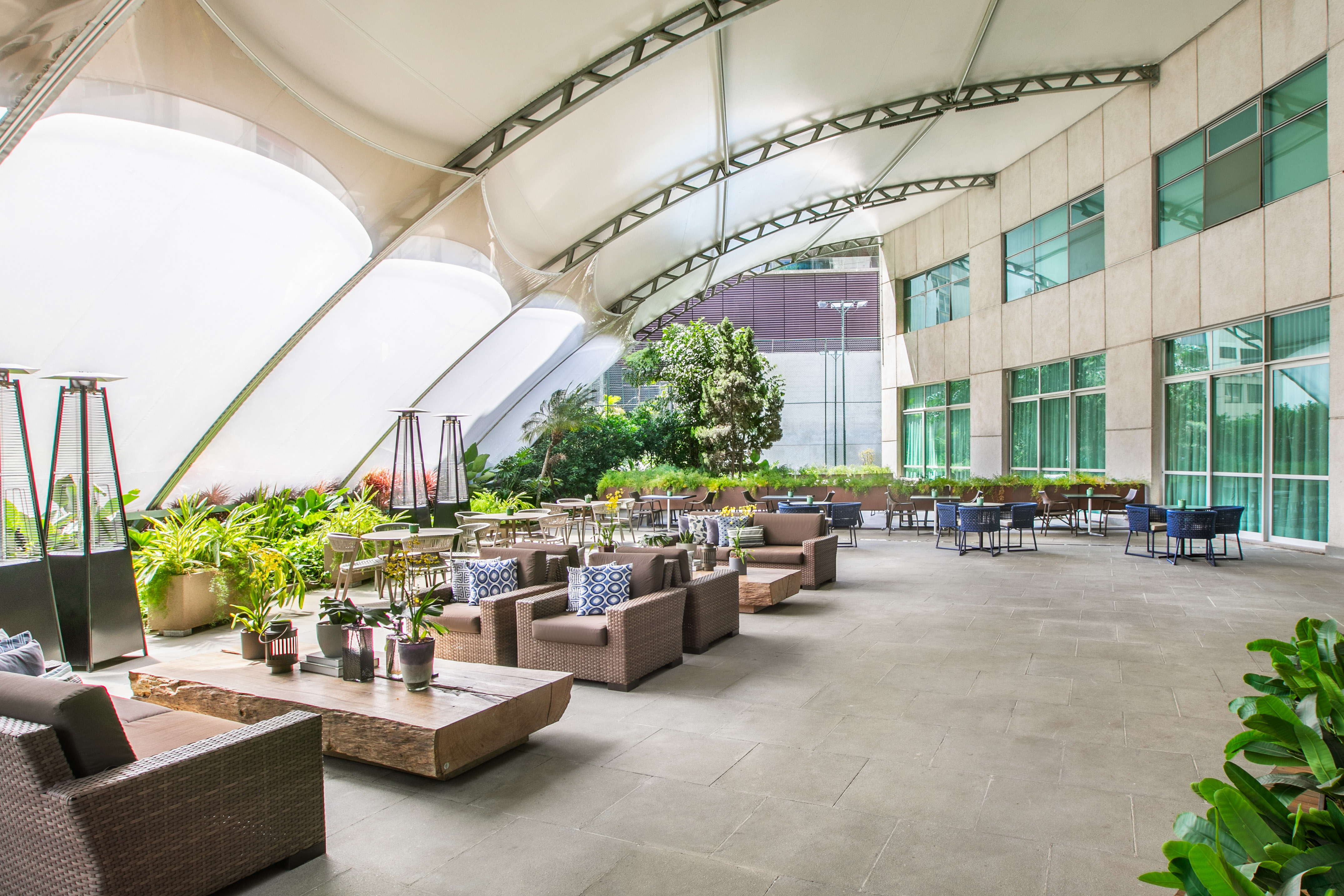 Modern outdoor patio lounge with canopy, diverse seating, lush plants, and dining tables. - Varanda - Meliá Jardim Europa, Sao Paulo