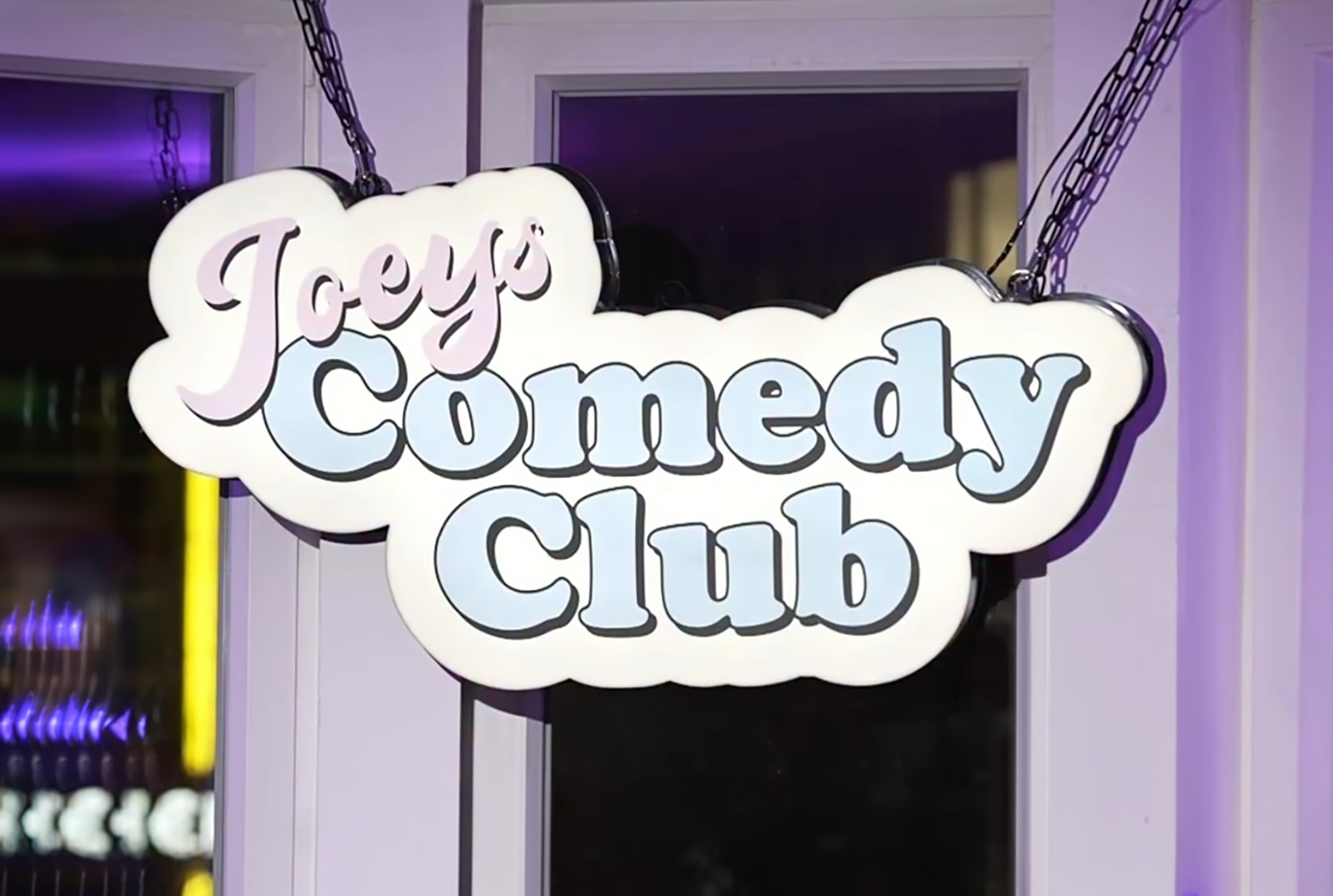 Hanging cloud-shaped sign for Joey's Comedy Club with pink and blue lettering.