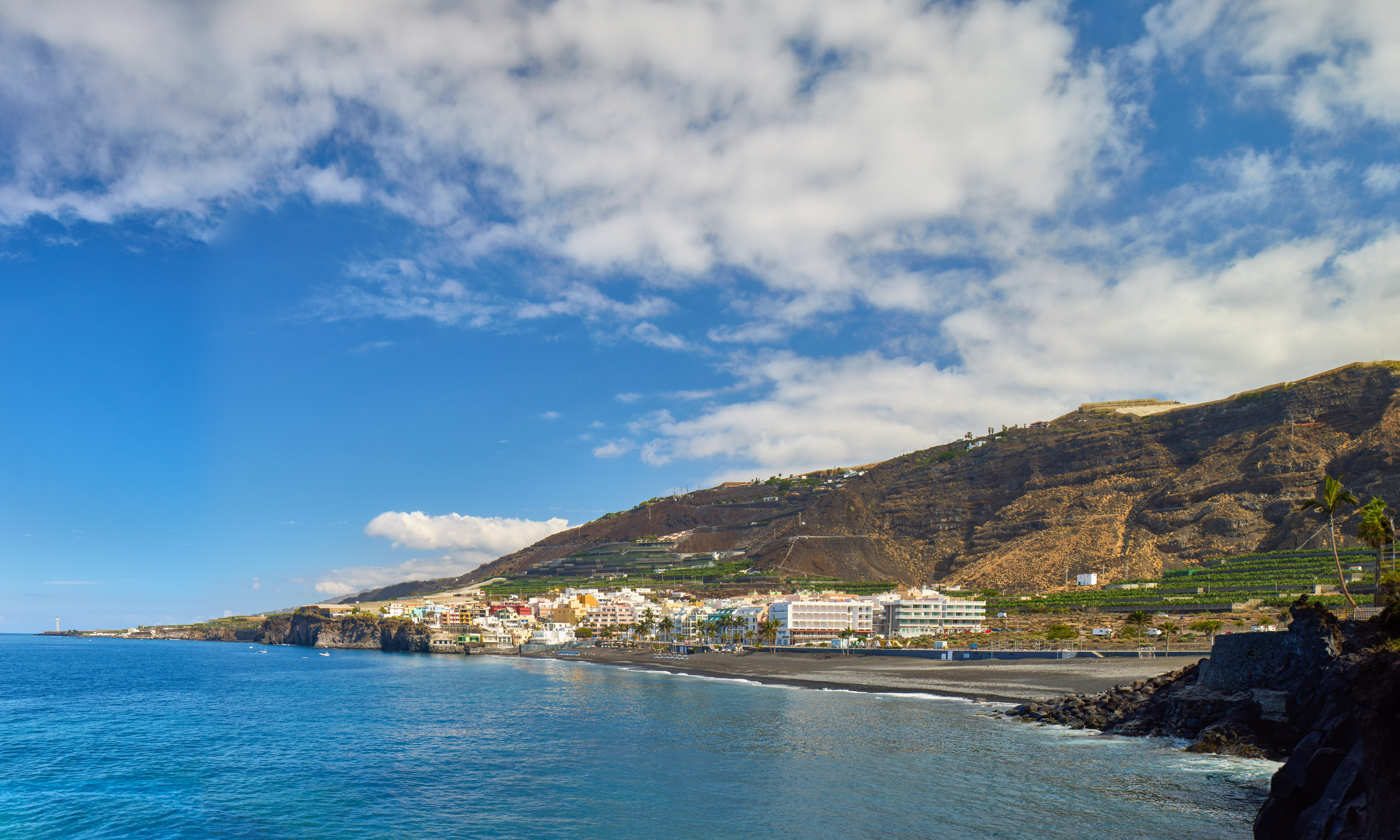 Coastal town with black sand beach, blue ocean, and steep volcanic hillside.