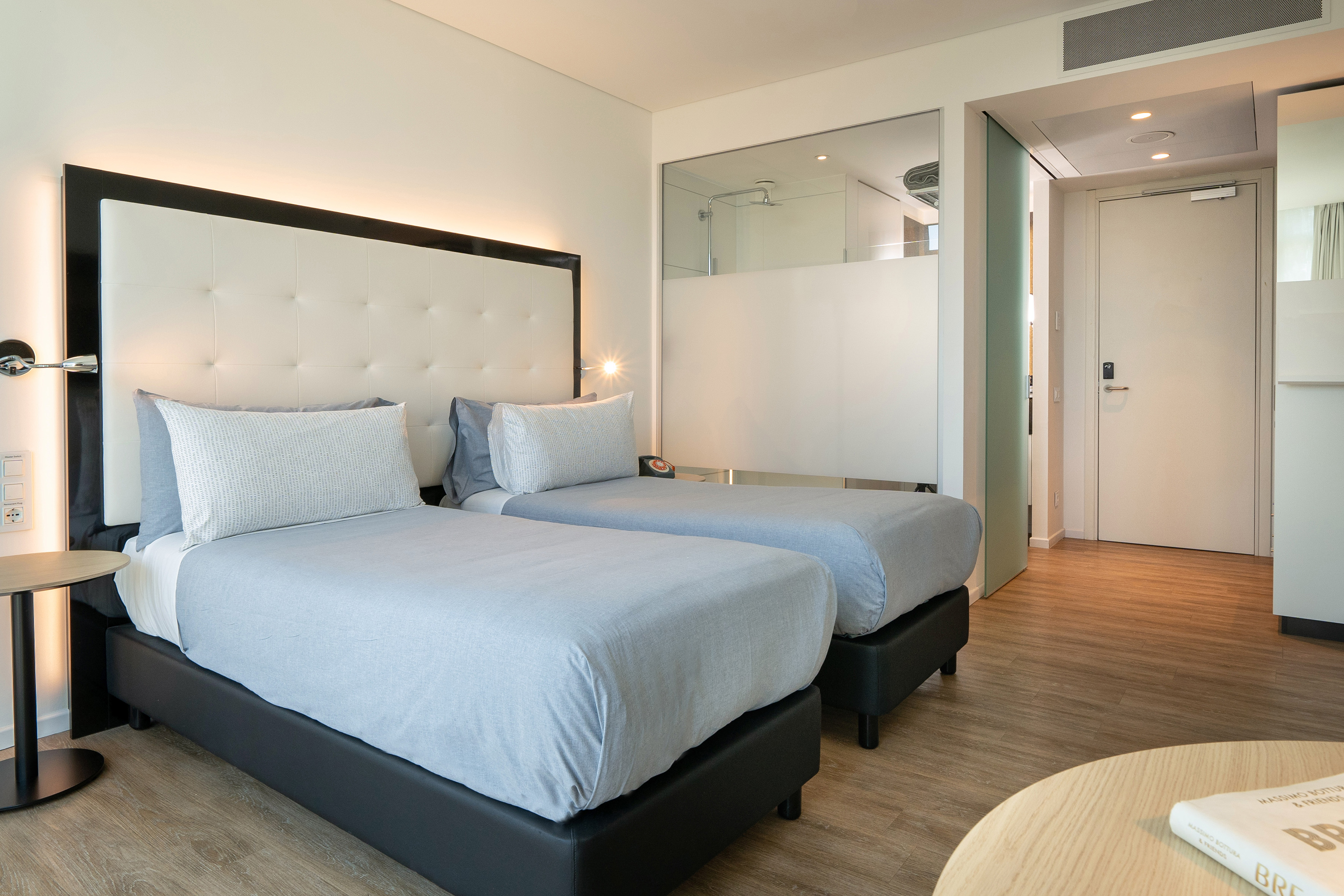 Modern hotel room with twin beds, light blue bedding, and minimalist decor.