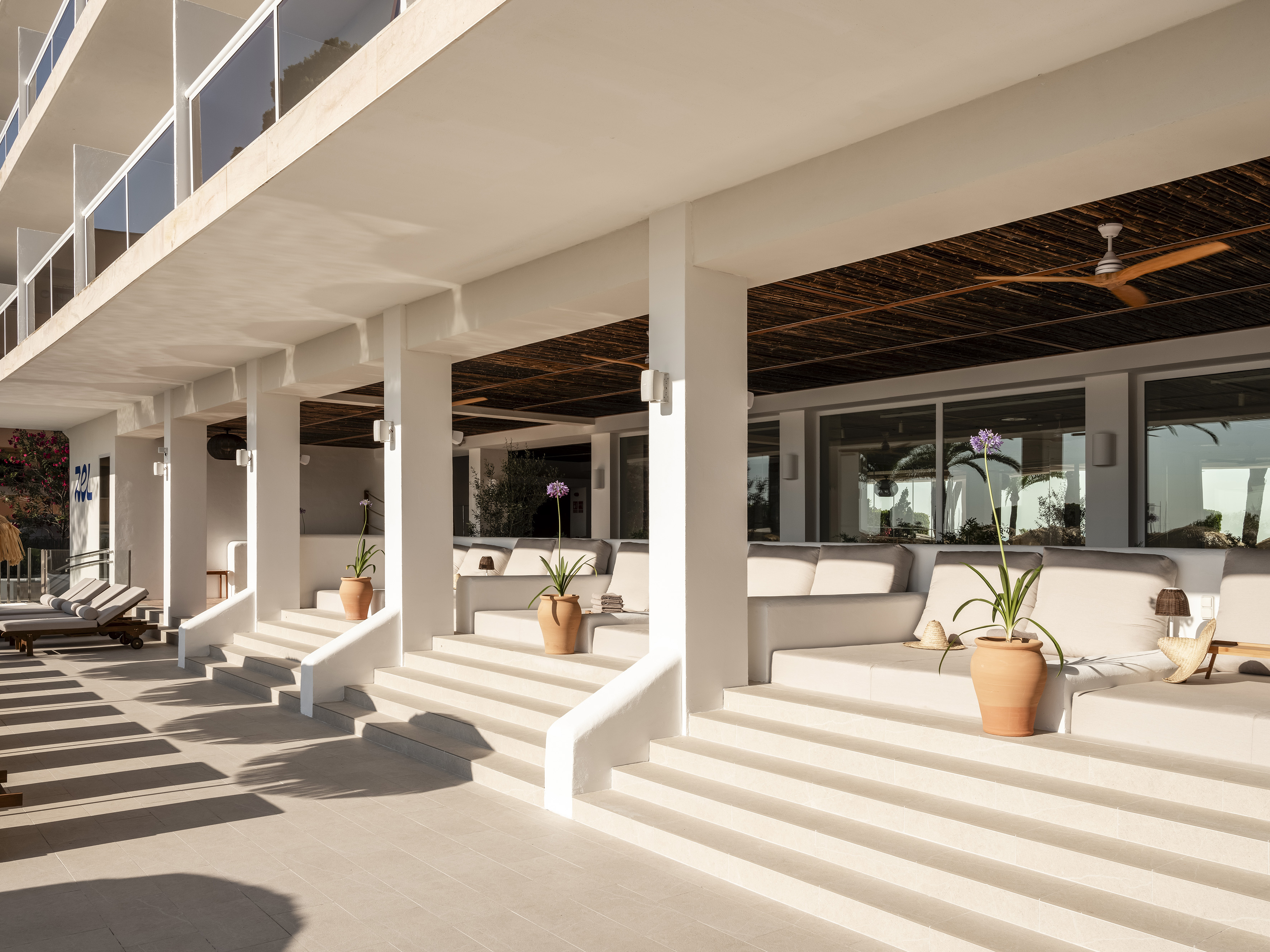 Sunny modern white hotel terrace with steps, daybeds, and potted plants.