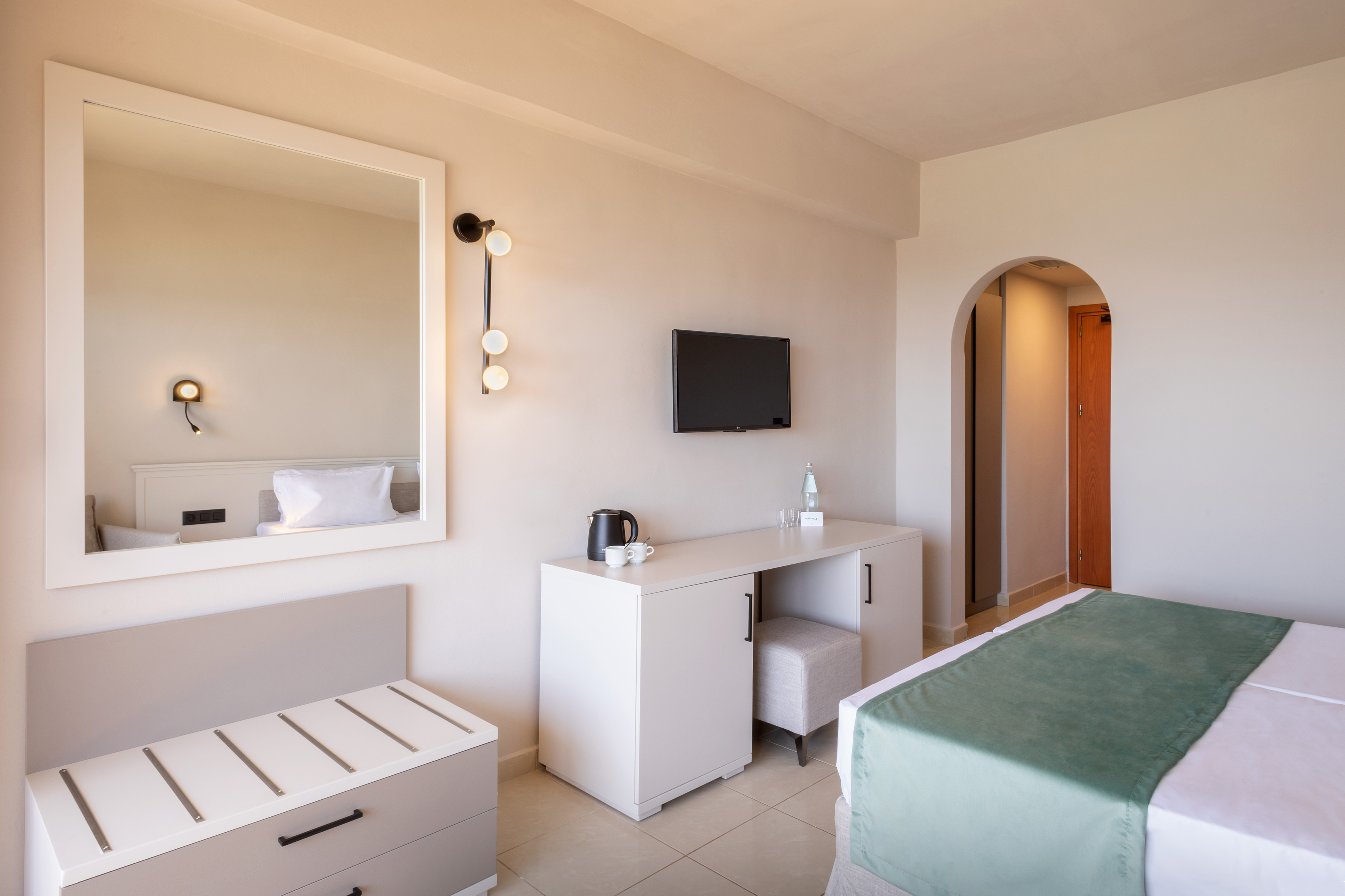 Modern hotel room with white furnishings, bed, TV, desk, and arched entry.