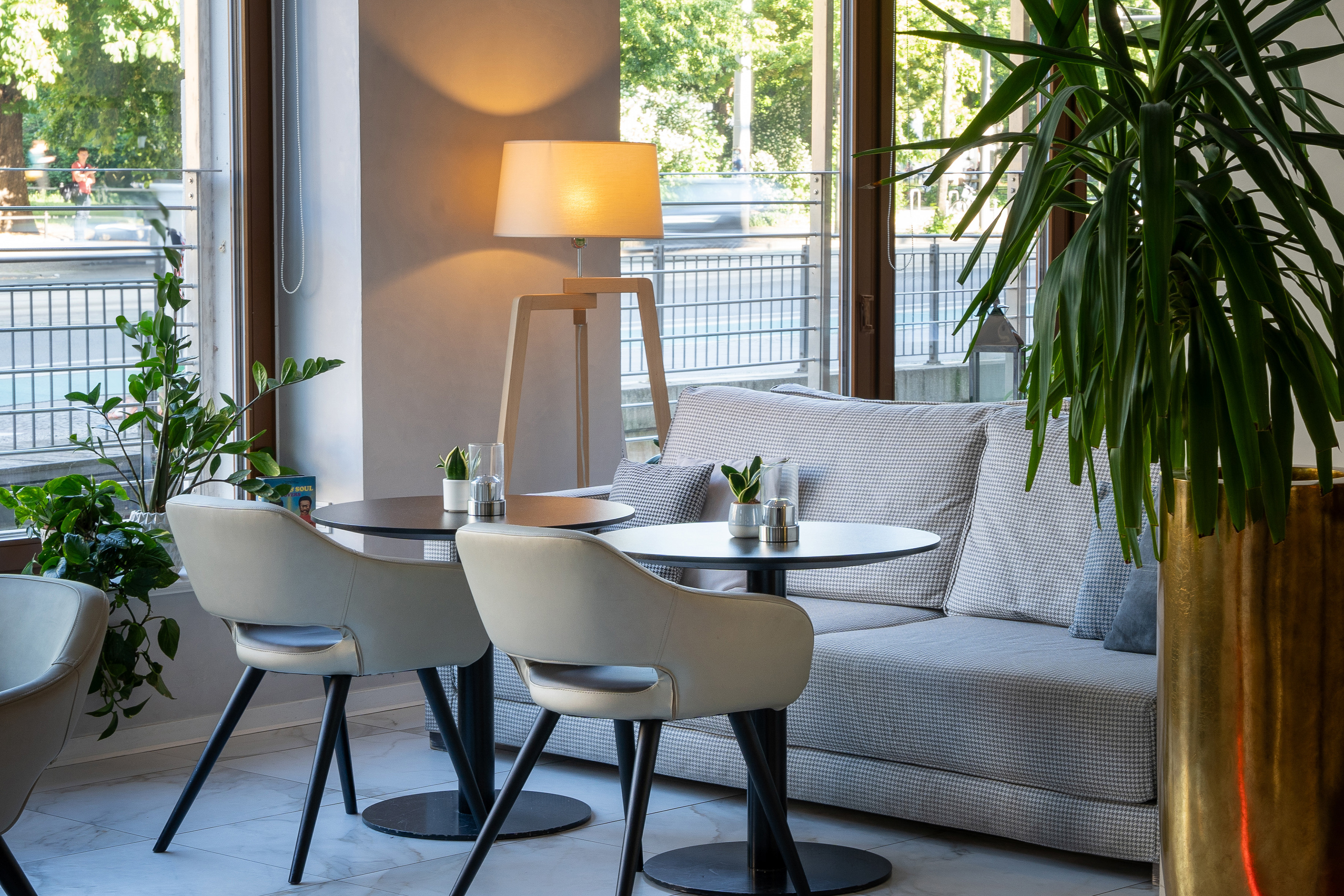Modern cafe lounge with sofa, chairs, tables, plants, and large windows.