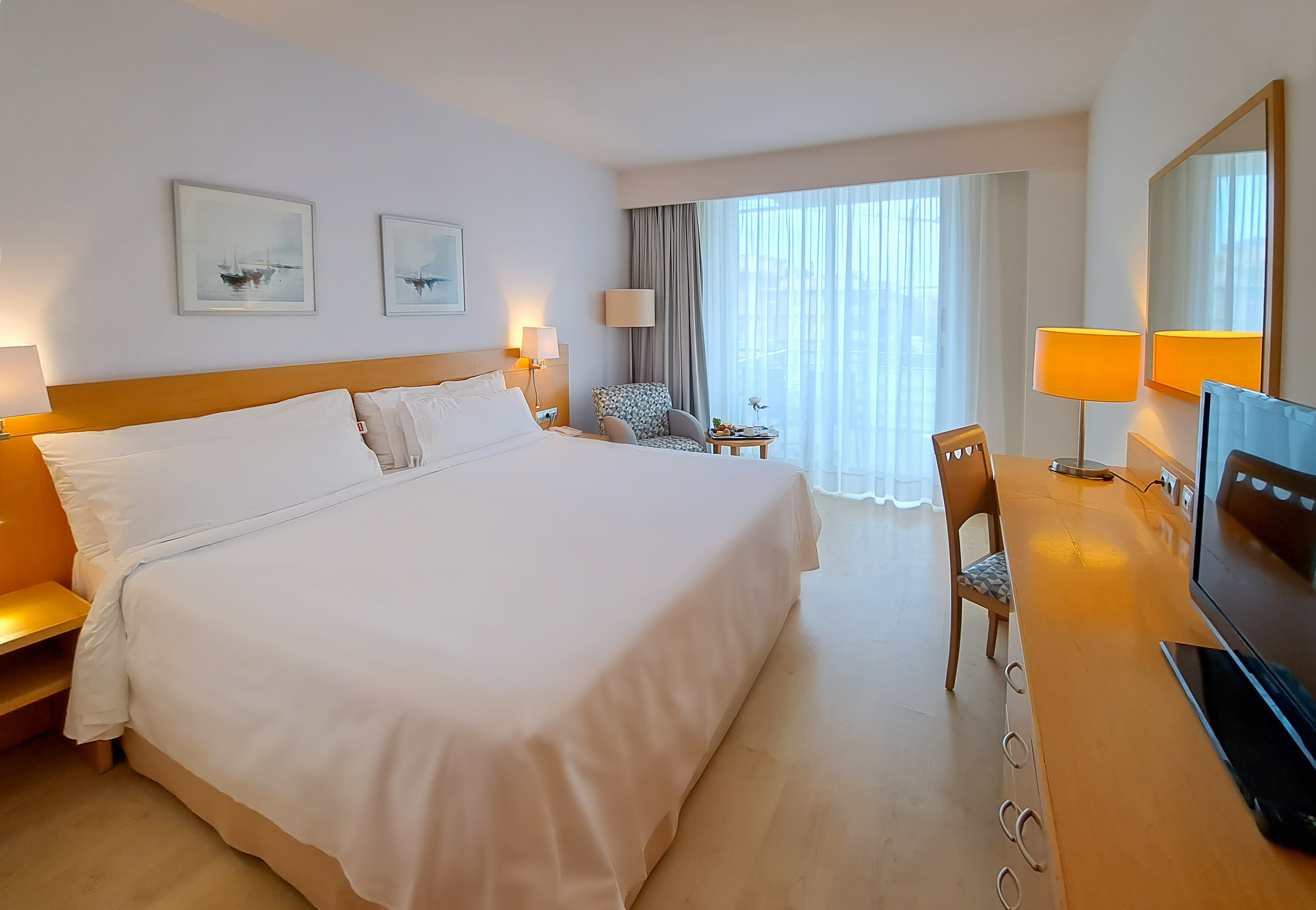 Spacious modern hotel room with king bed, desk, armchair, and bright window.