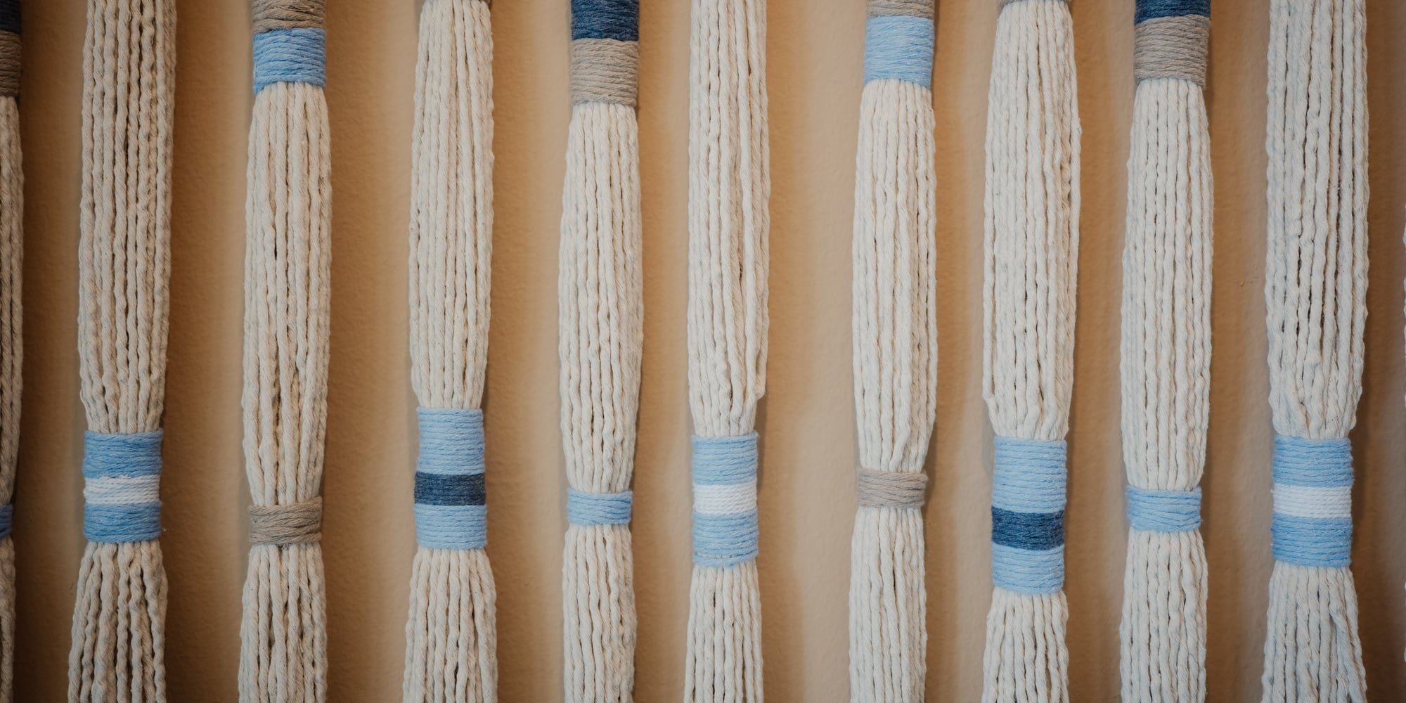 Arte de pared macramé con hilos crema, bandas azules y grises.
