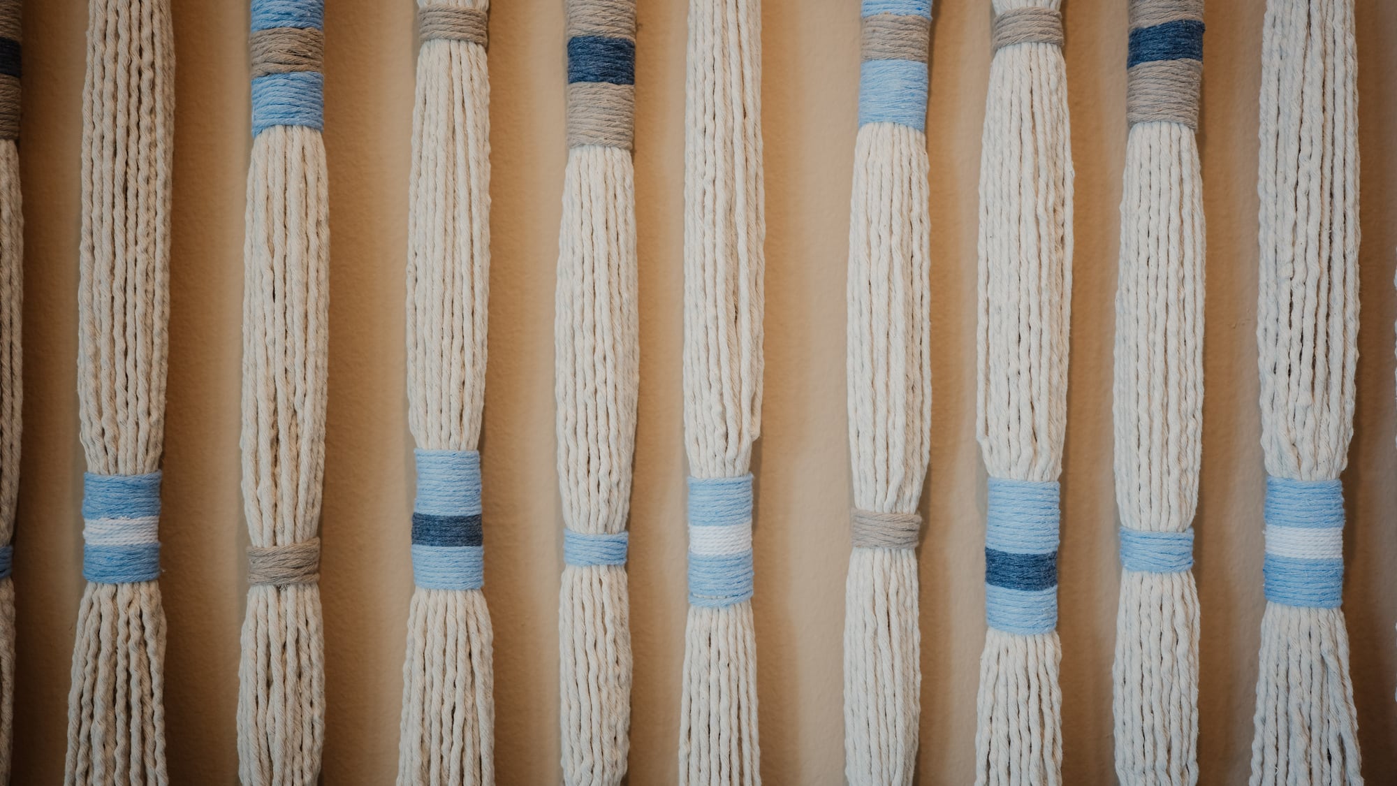 Arte de pared macramé con hilos crema, bandas azules y grises.