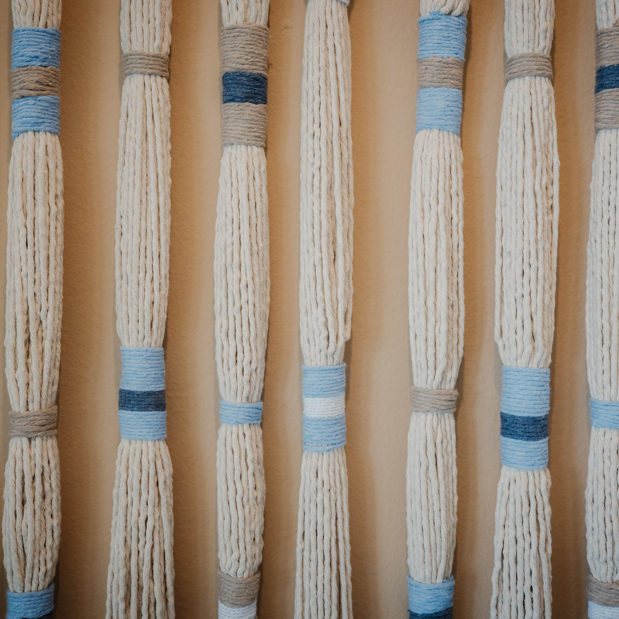 Arte de pared macramé con hilos crema, bandas azules y grises.