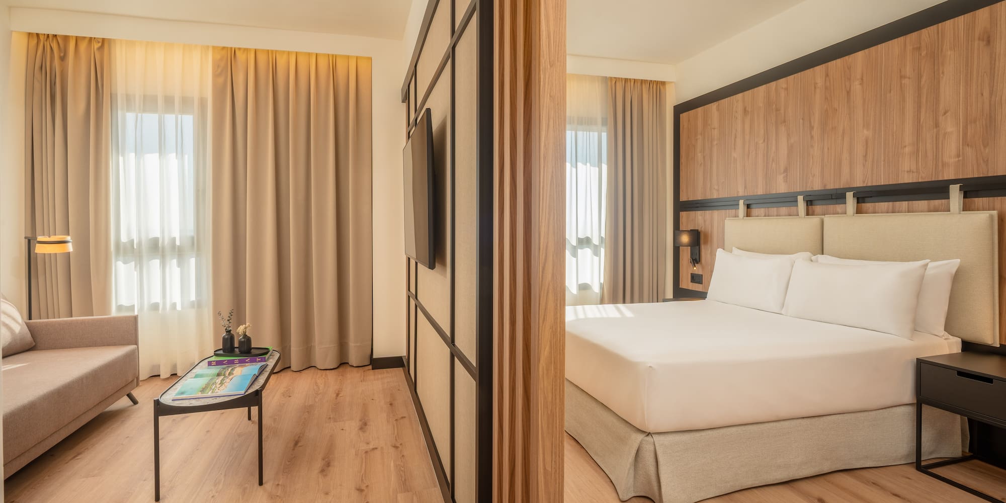 Modern hotel suite with separate living and bedroom, wood floor, neutral tones.