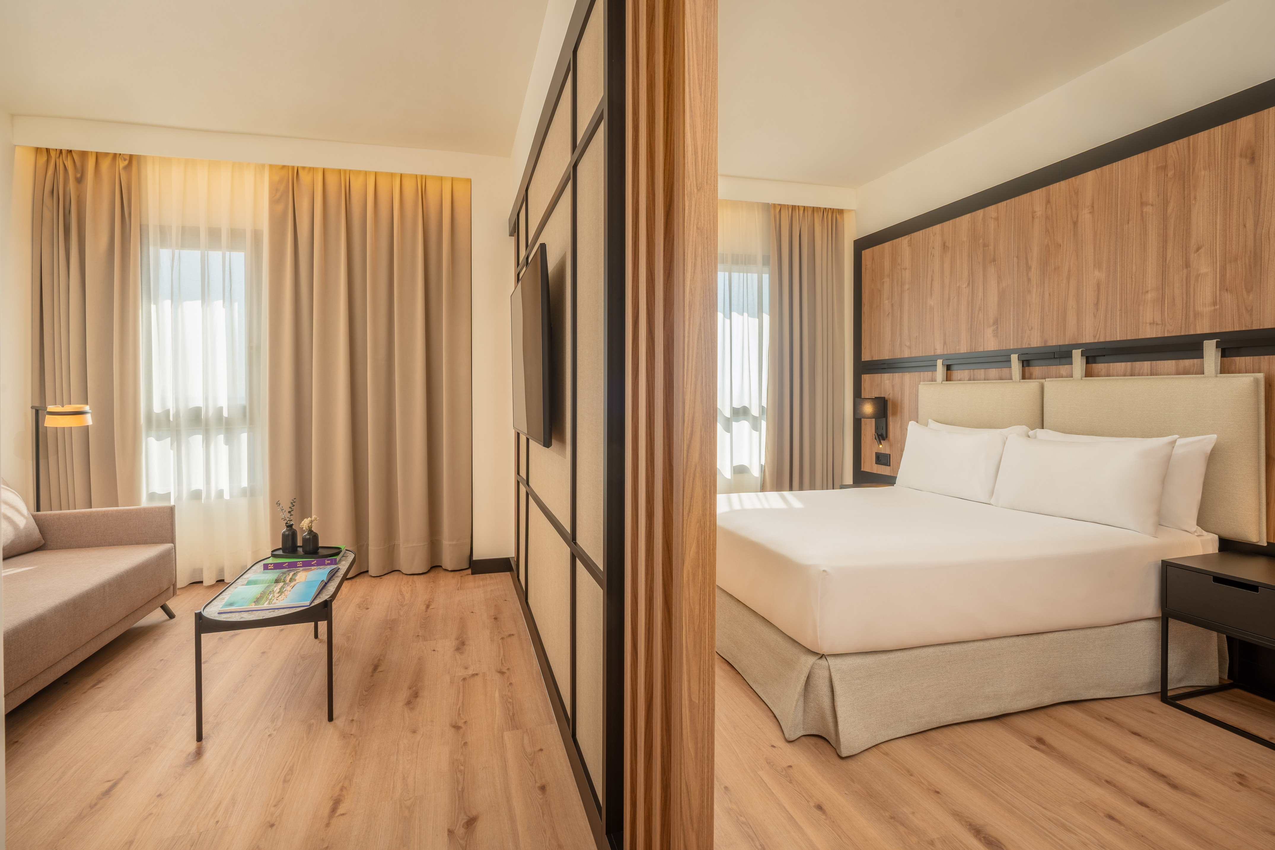 Modern hotel suite with separate living and bedroom, wood floor, neutral tones.