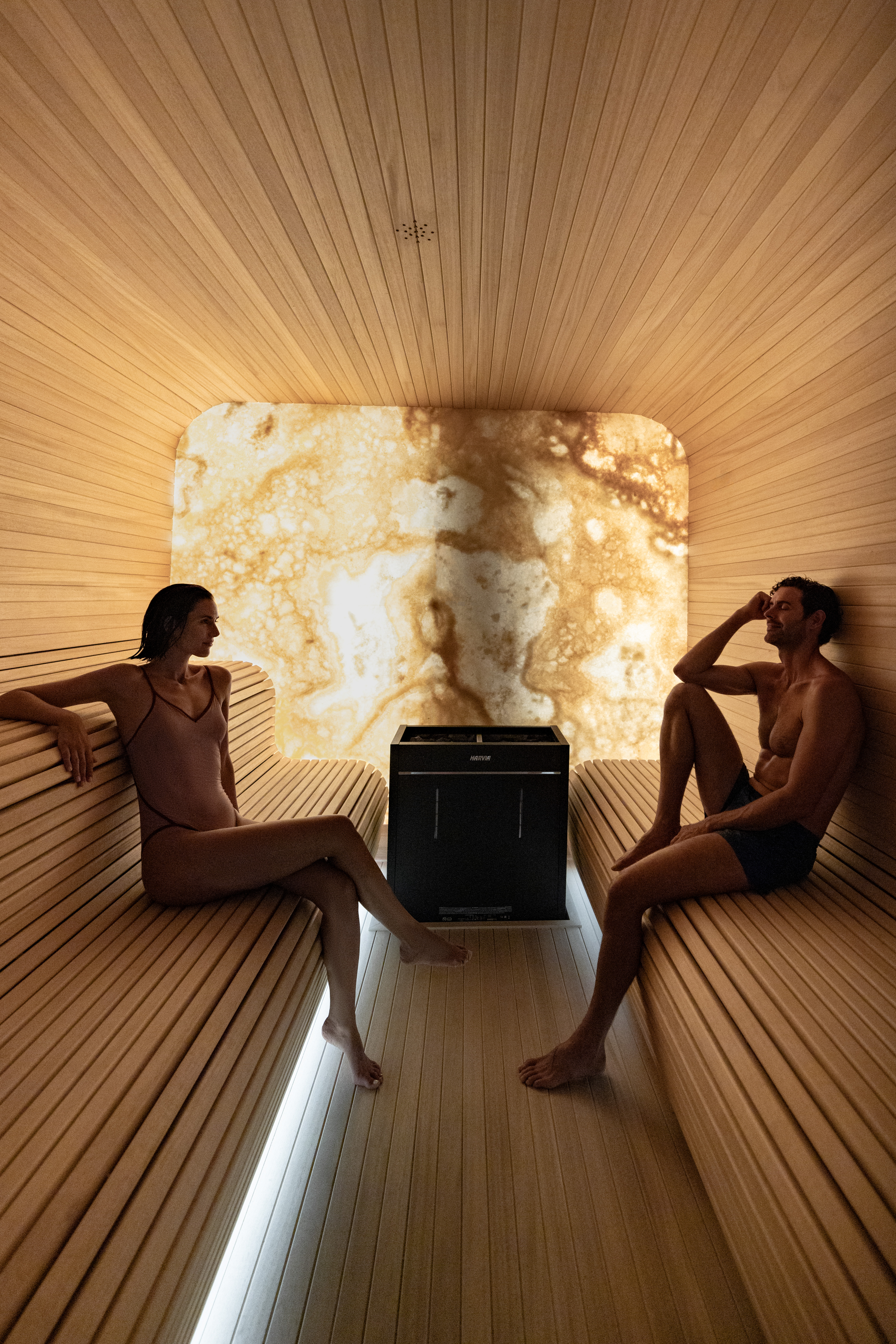 Modern wooden sauna with man, woman, Harvia heater, and glowing back wall.