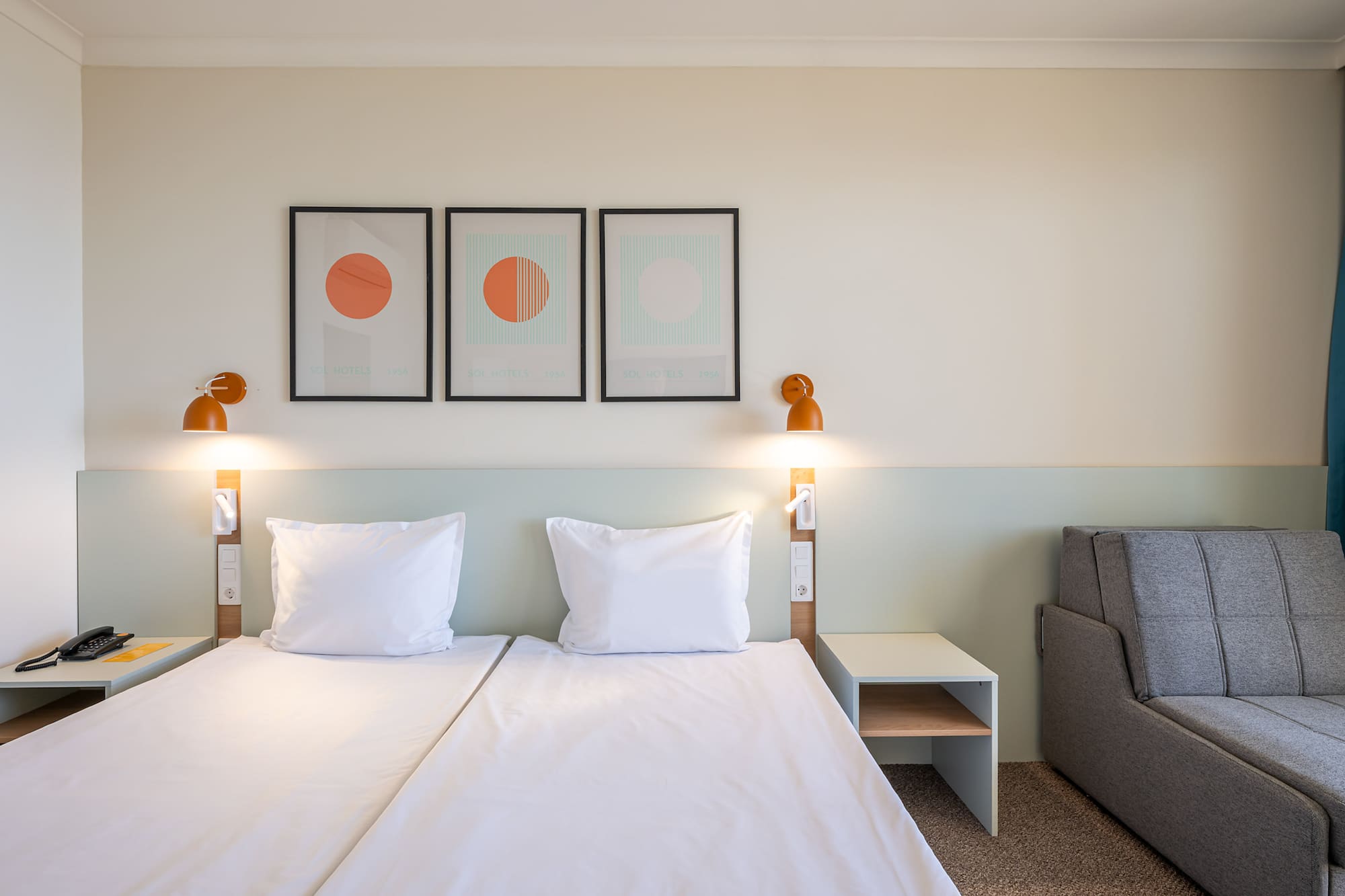 Modern hotel room with twin beds, orange lights, geometric art, and sofa.