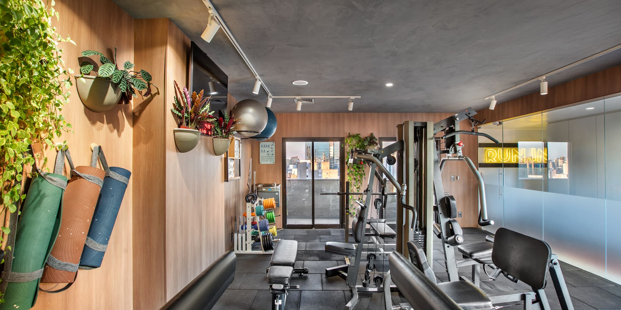 Modern fitness room with workout machines, weights, yoga mats, city skyline view.