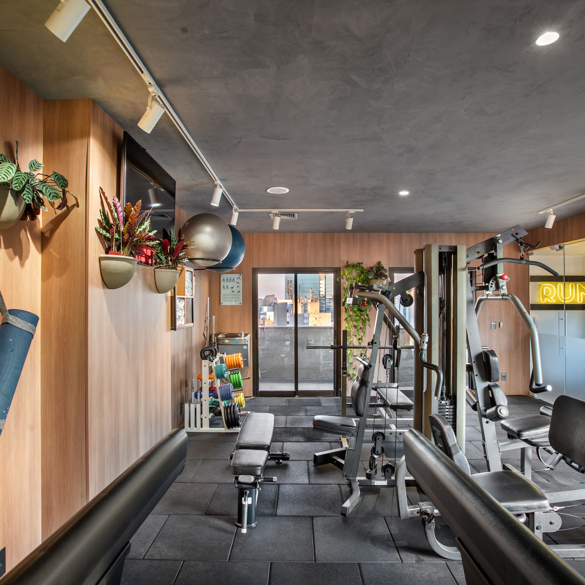 Modern fitness room with workout machines, weights, yoga mats, city skyline view.