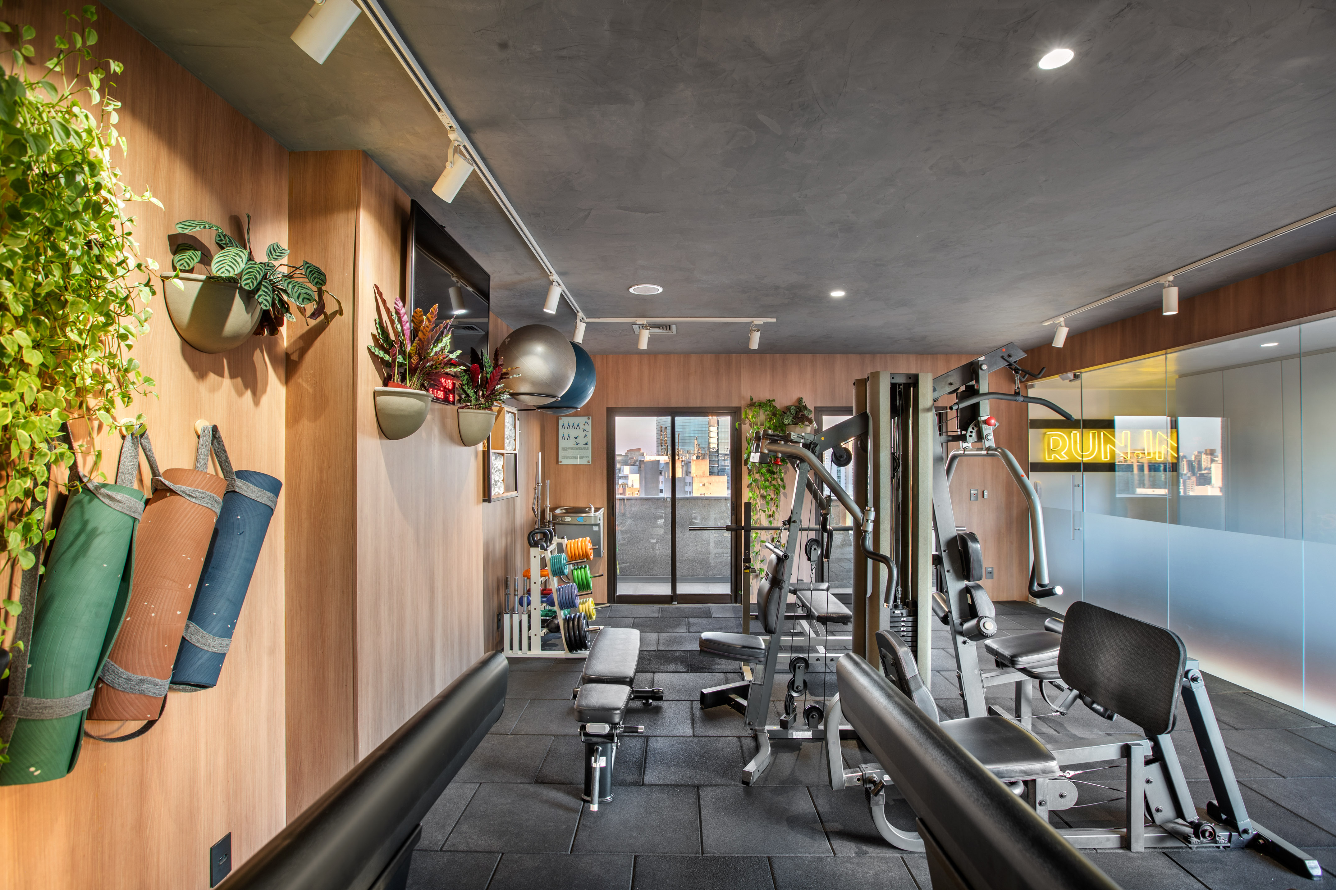 Modern fitness room with workout machines, weights, yoga mats, city skyline view.