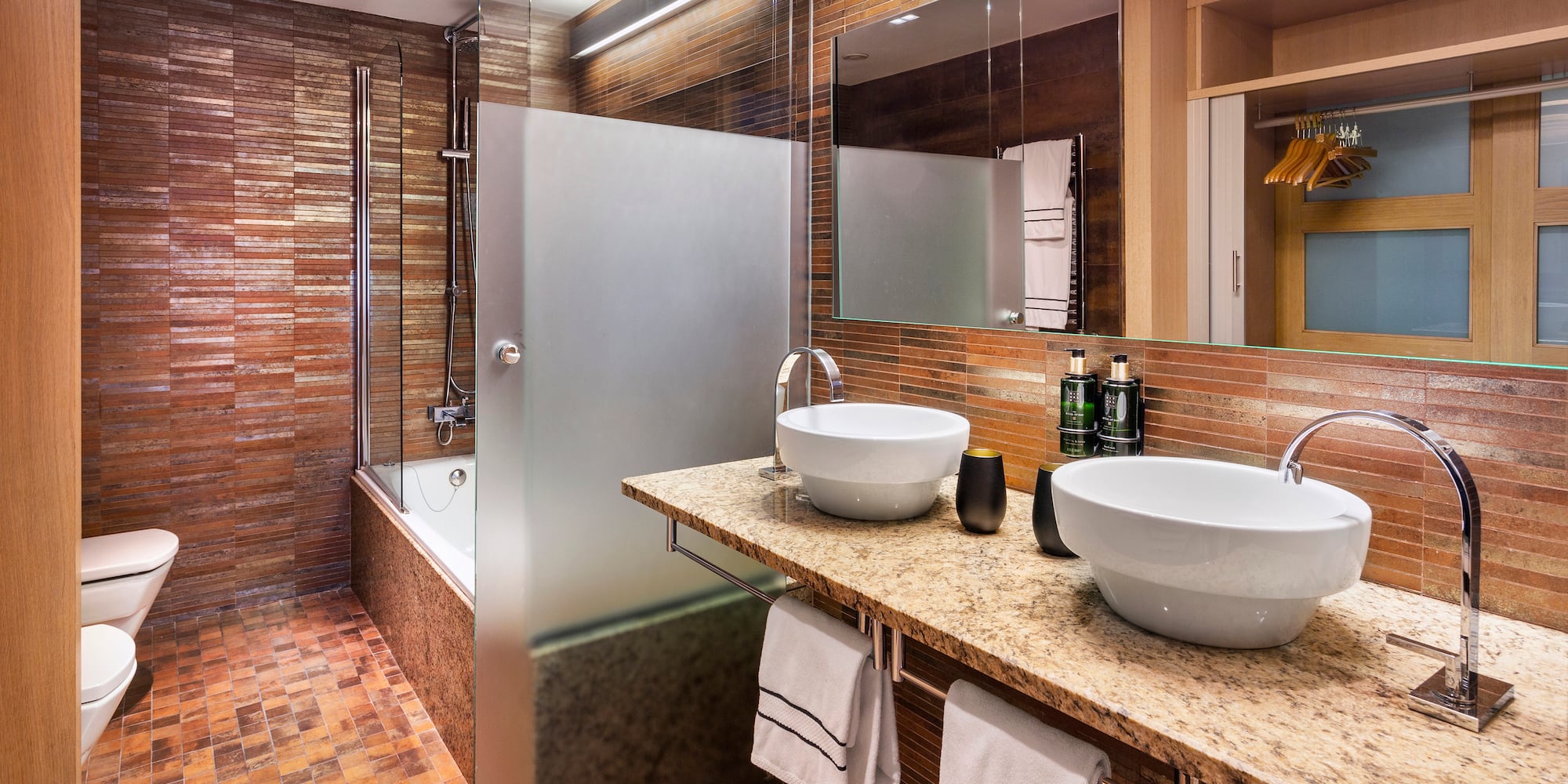 Baño moderno con lavabos dobles, encimera granito y azulejos marrones. Ducha y bañera.