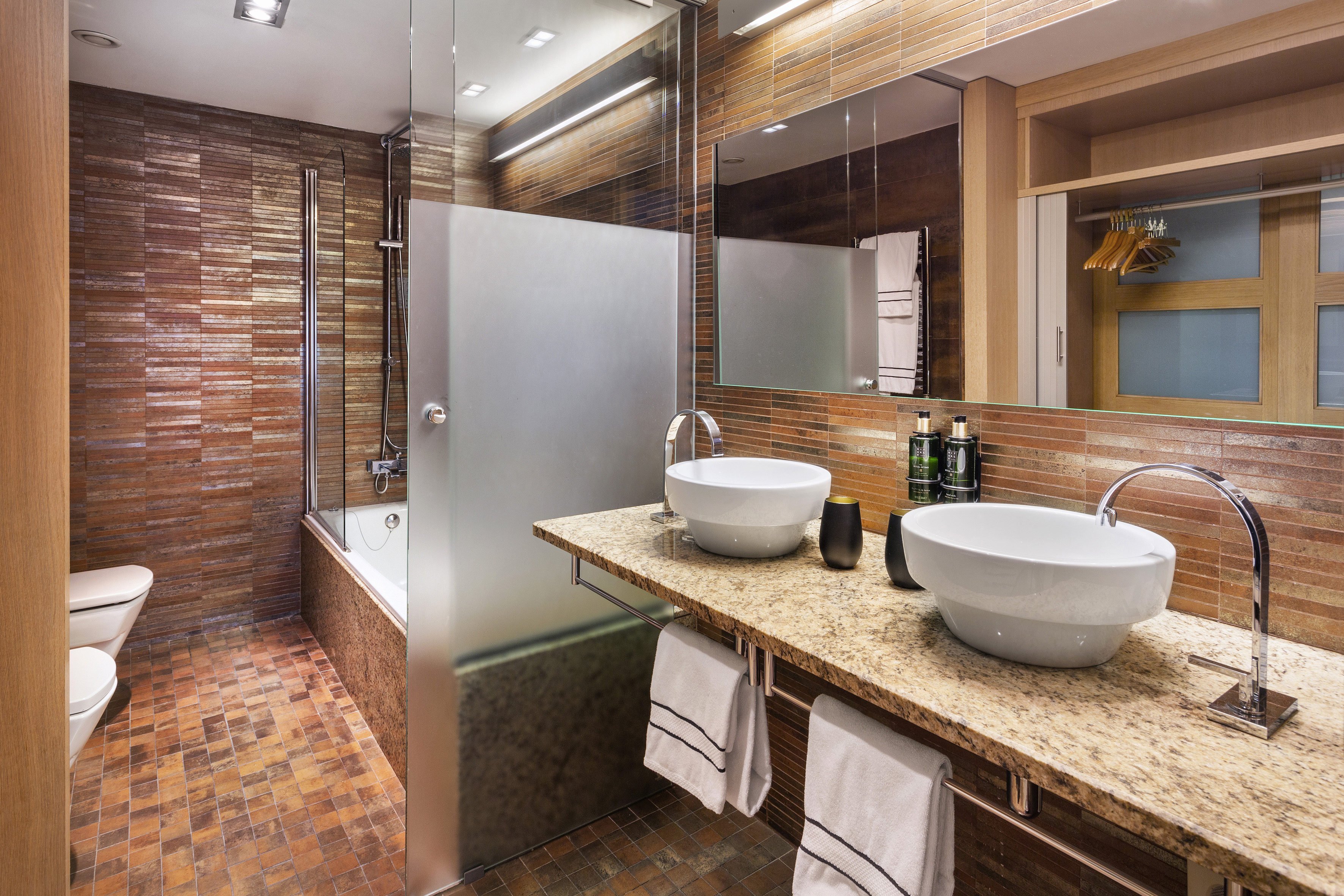 Baño moderno con lavabos dobles, encimera granito y azulejos marrones. Ducha y bañera.