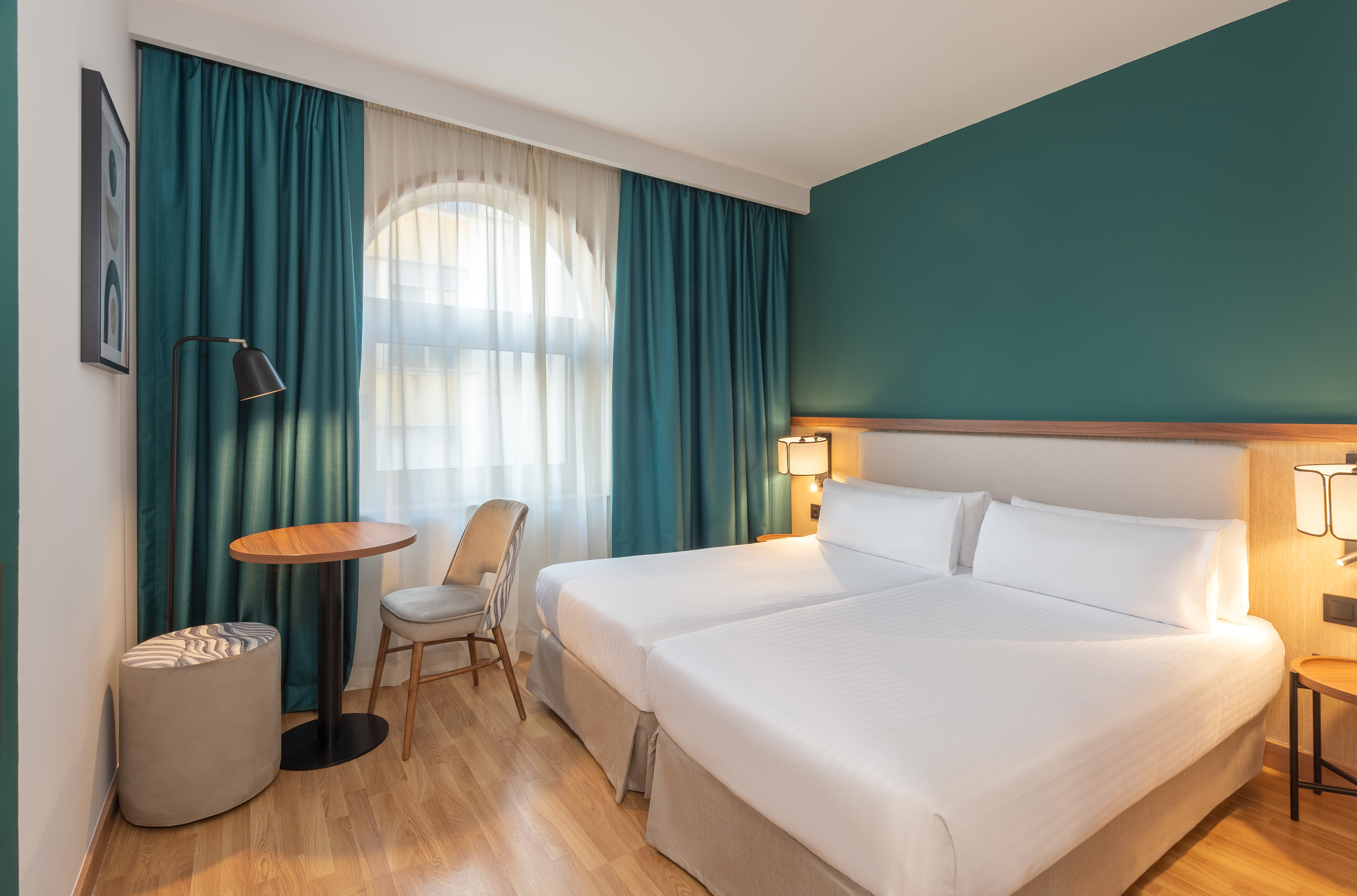 Modern hotel room with two twin beds, teal accent wall, and arched window.
