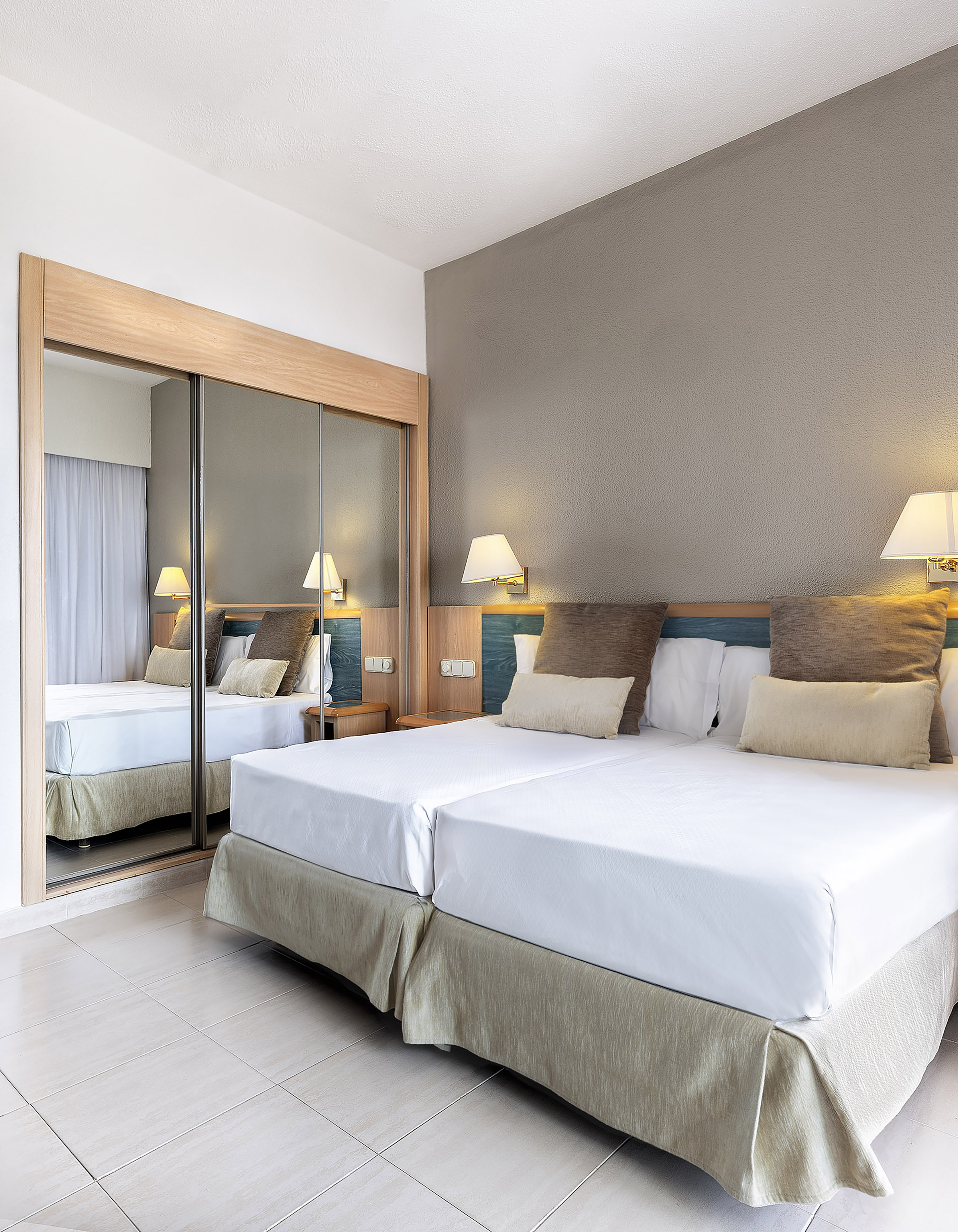 Hotel bedroom with twin beds, wooden headboard, wall lamps, and mirrored wardrobe.