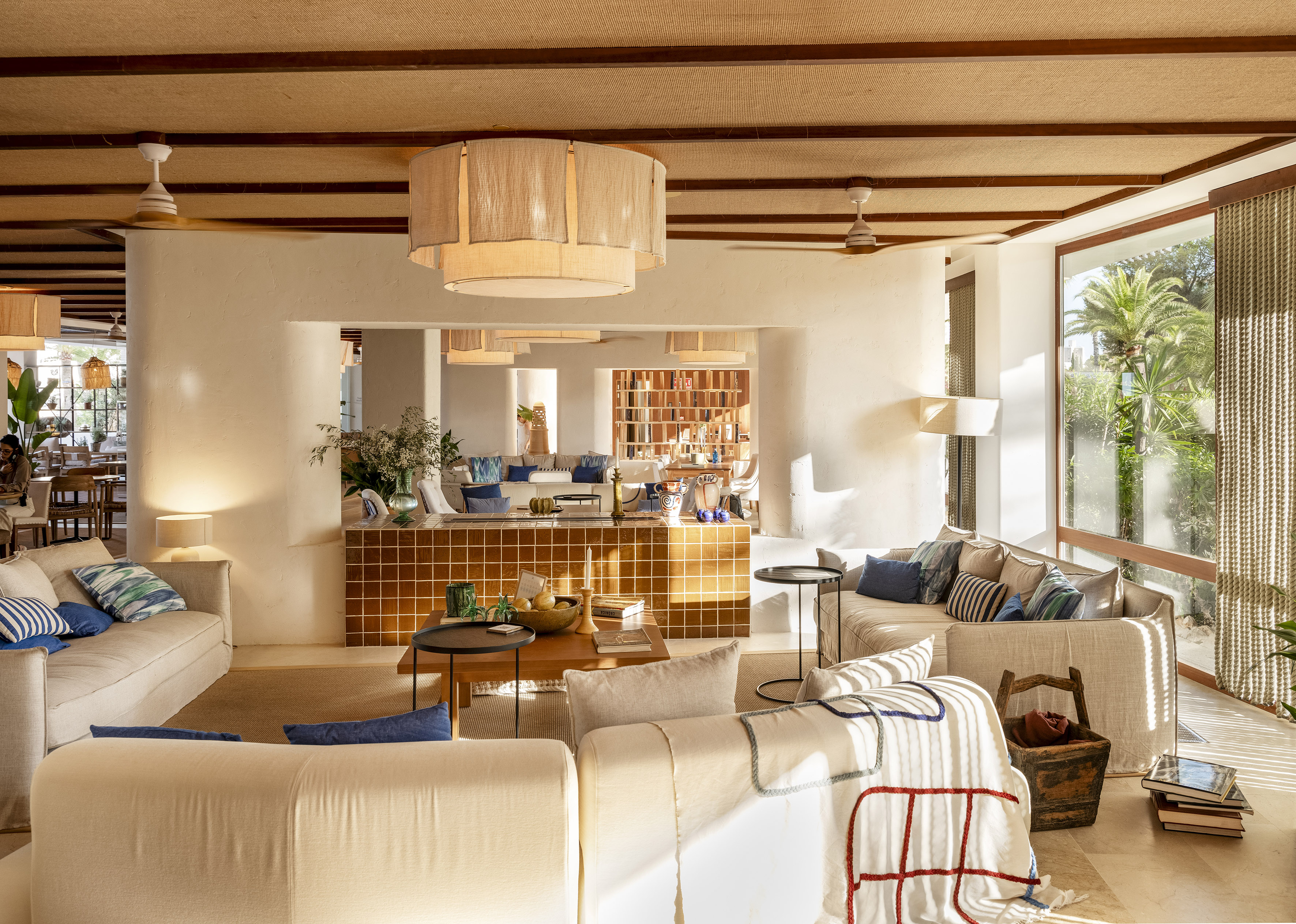 Sunny Mediterranean lounge with white sofas, woven ceiling, natural light, palm trees.