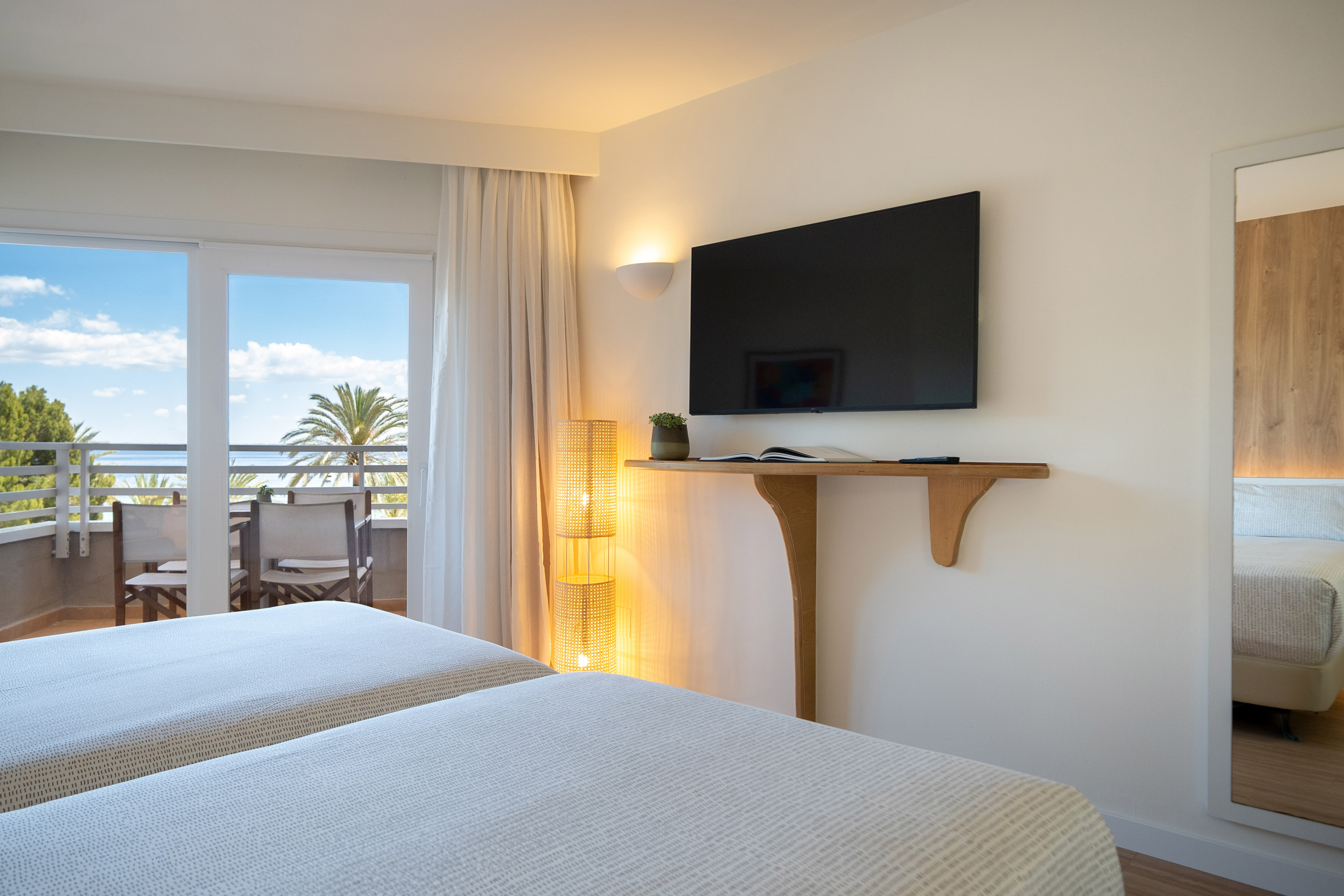 Bright hotel room with twin beds, ocean view balcony, and wall-mounted TV.
