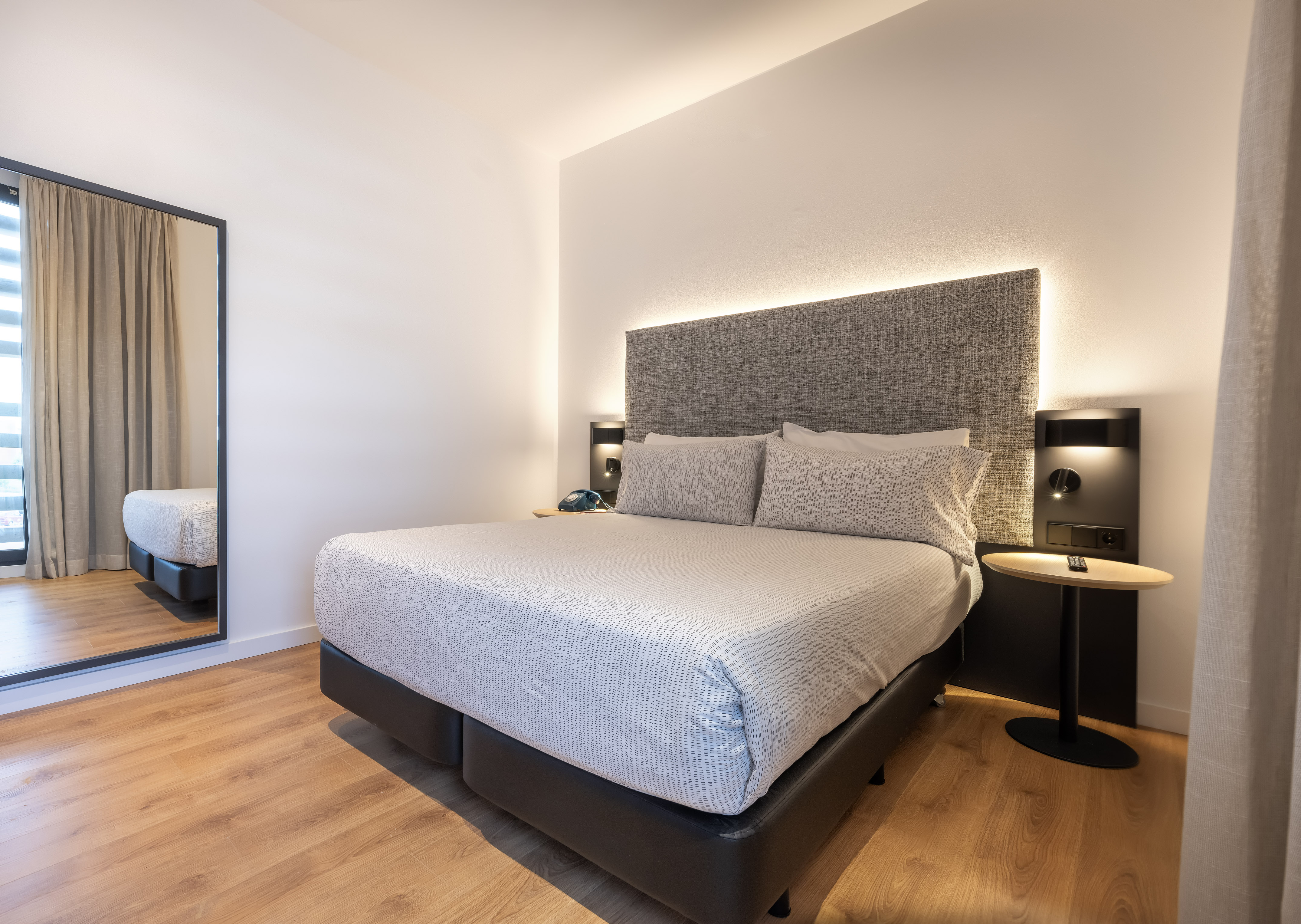 Modern hotel bedroom with large bed, backlit headboard, wood floor, and mirror.