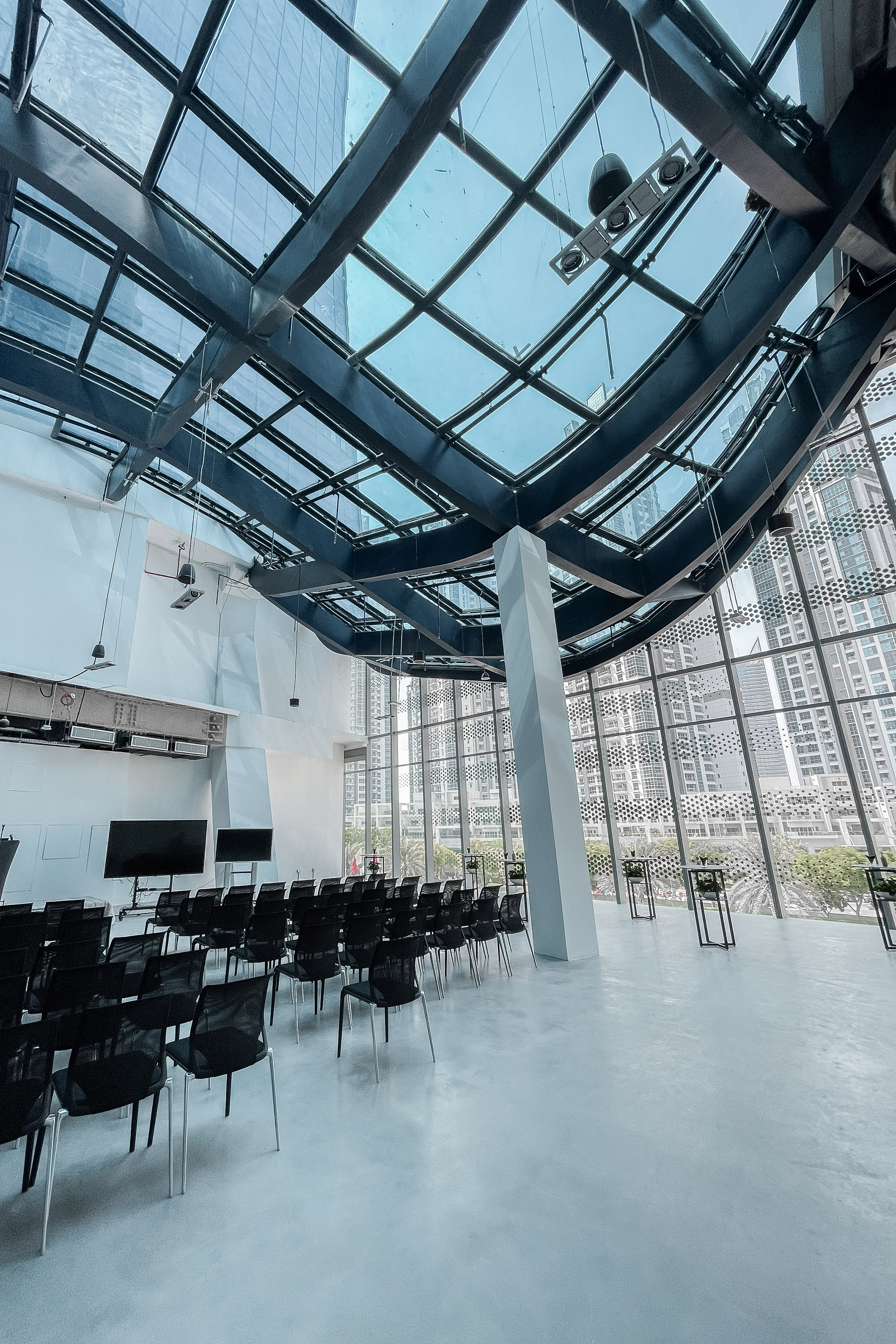Modern conference room with glass ceiling, city view, black chairs, and large screens.
