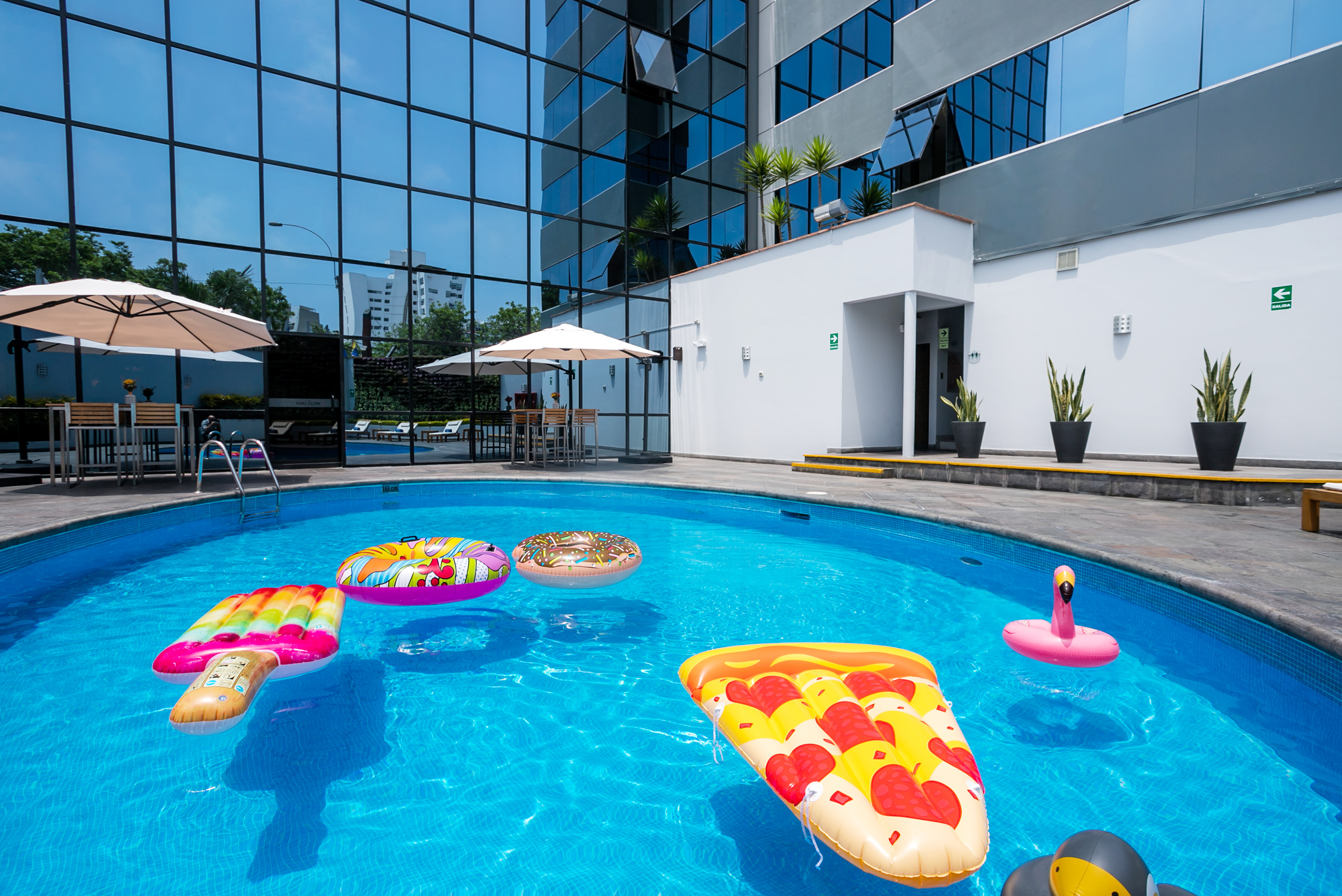 Vibrant hotel pool with pizza, flamingo floats, and modern glass building.