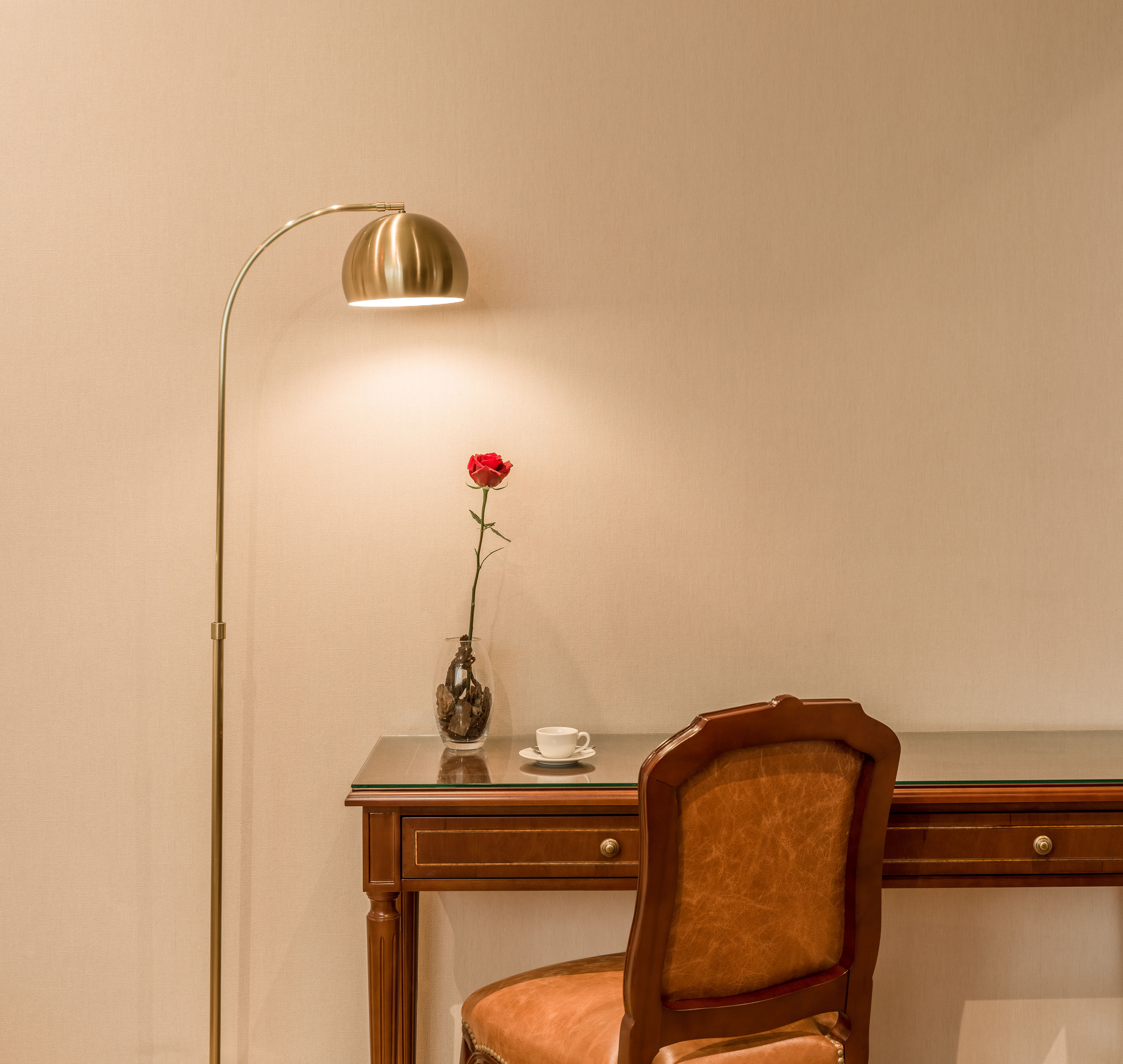 Hotel room desk with golden lamp, leather chair, red rose, and teacup.