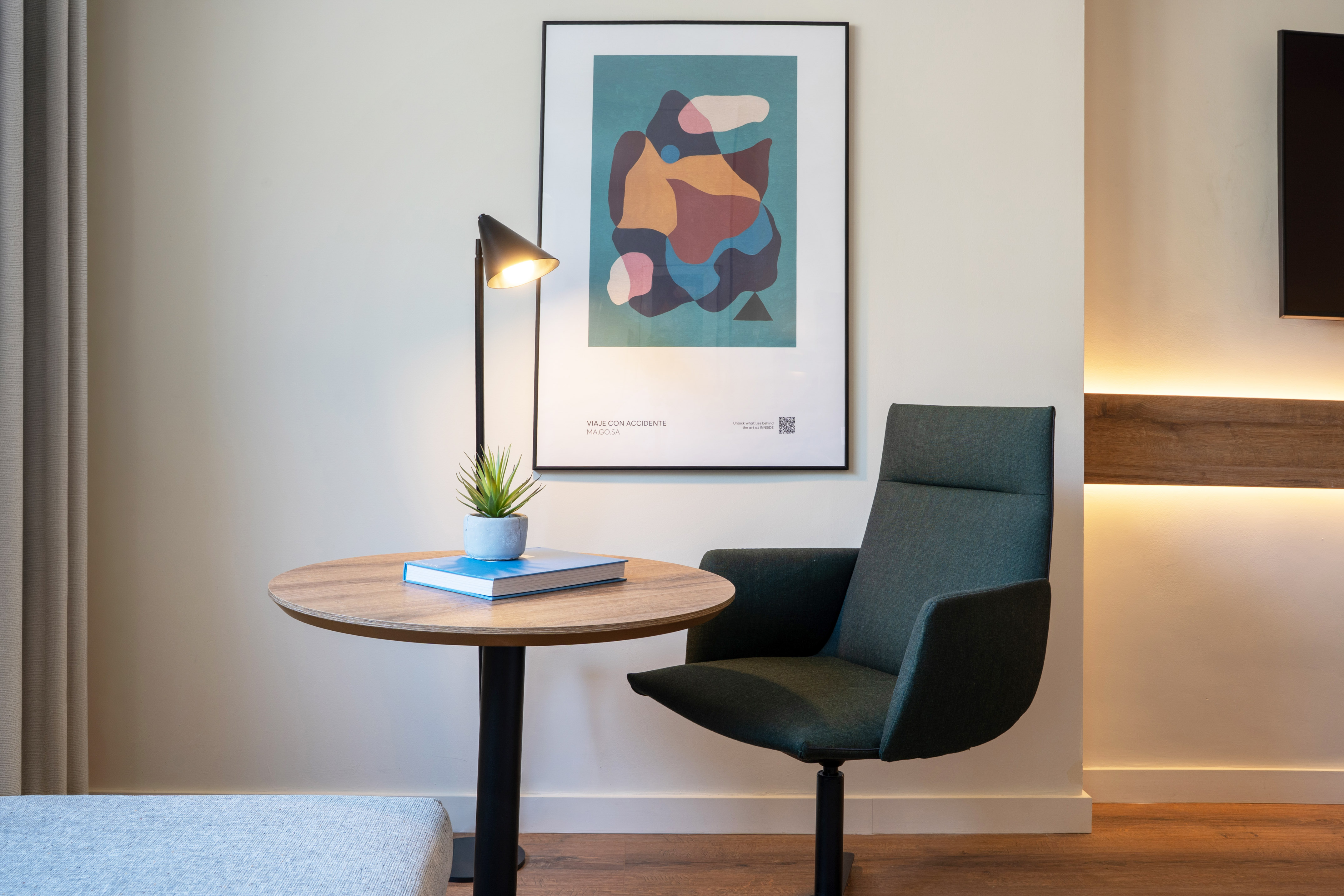 Modern hotel room with green armchair, round wood table, lamp, and abstract art.