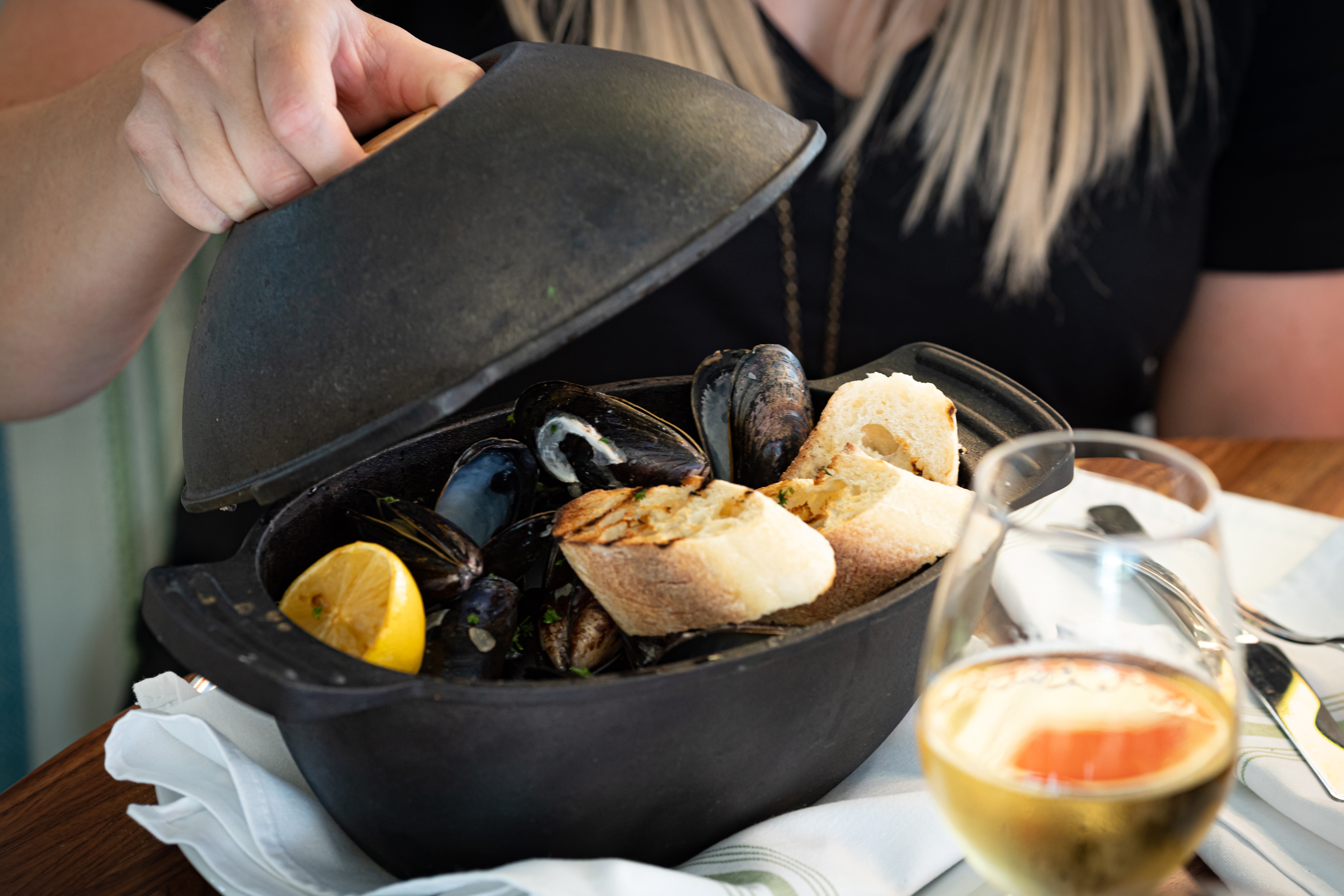 Hand opening cast iron pot of mussels, crusty bread, and lemon.