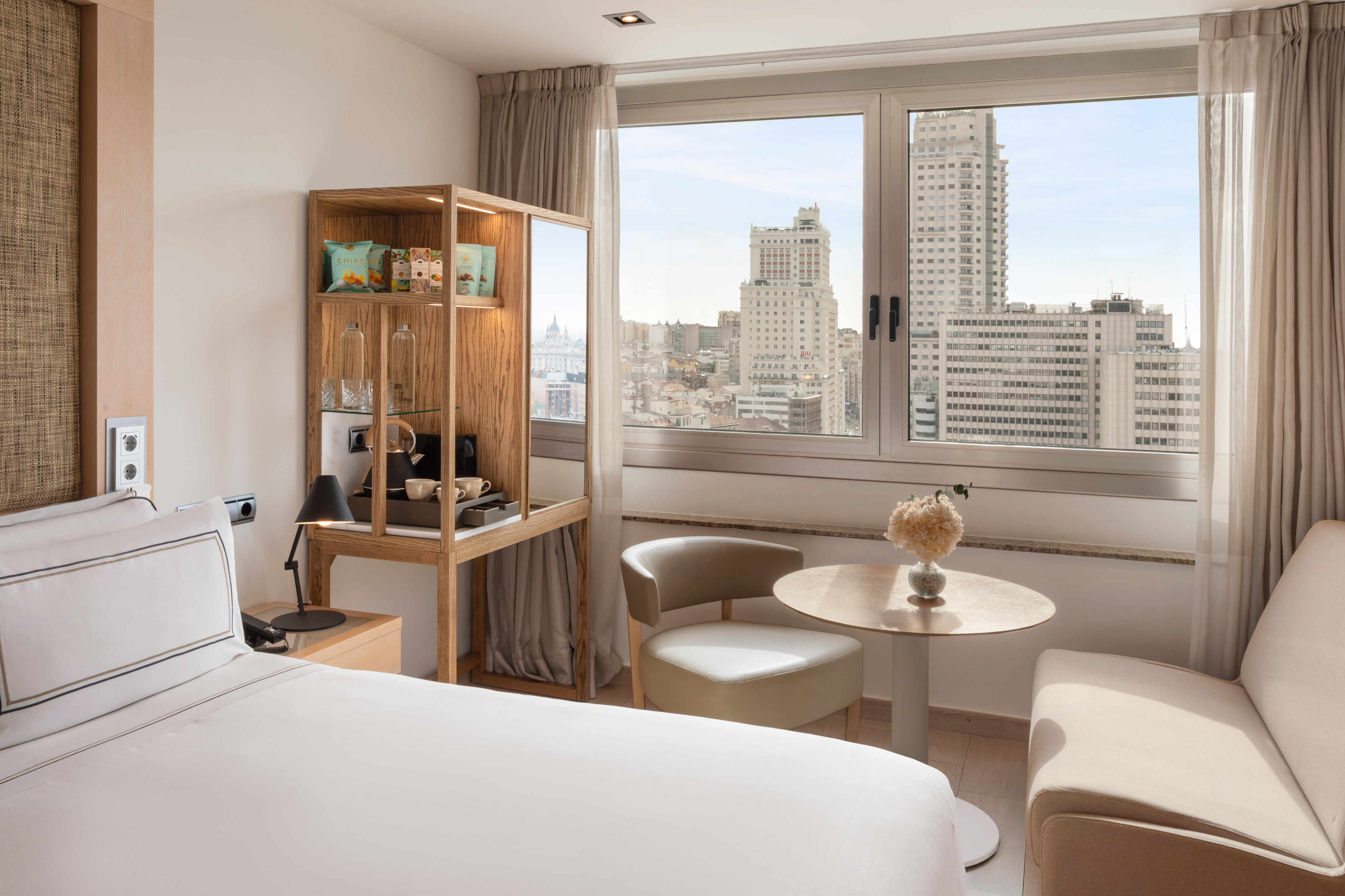 Bright hotel room with Madrid city view, bed, cozy seating, and minibar.