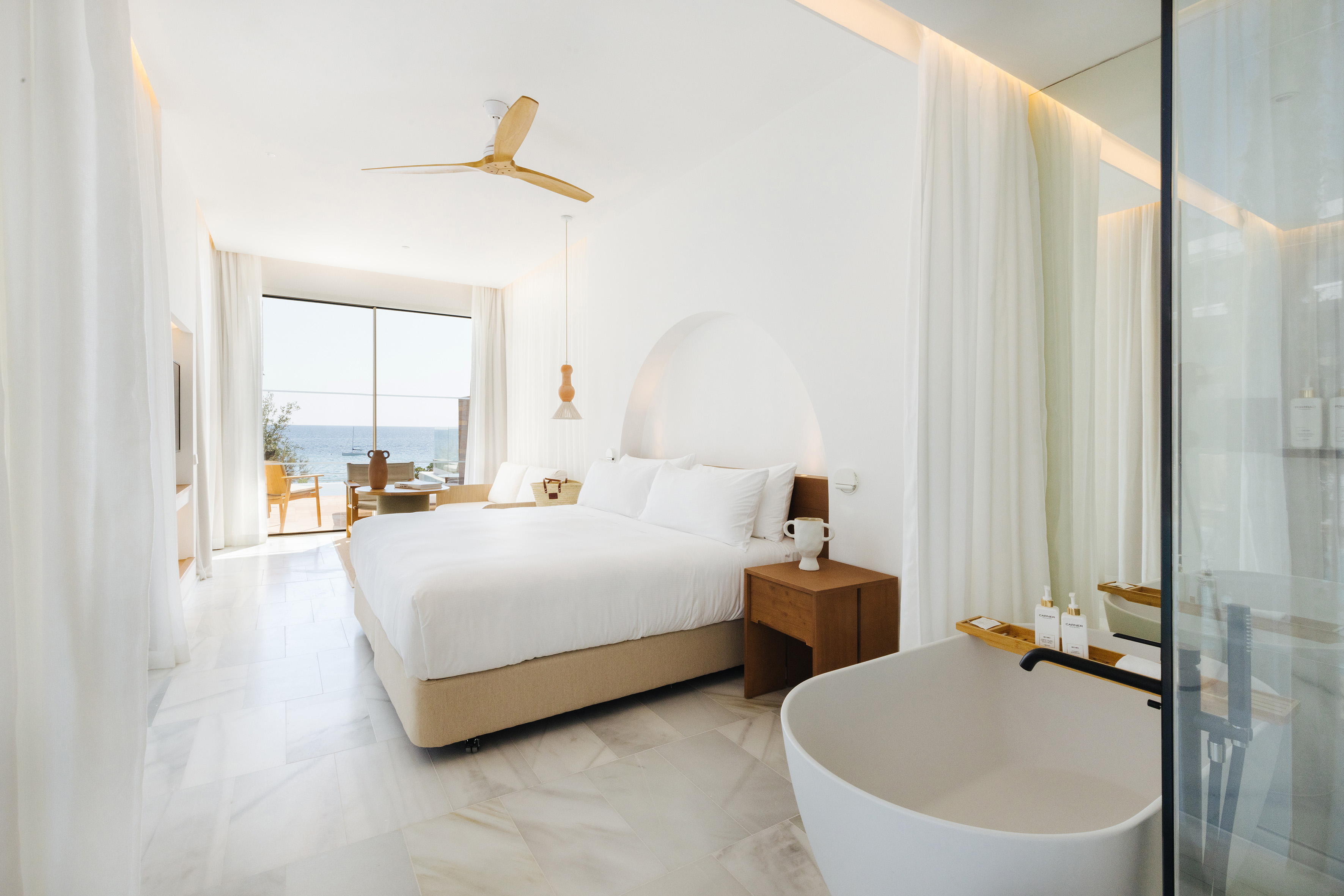 Modern white hotel room with king bed, ocean view, and freestanding bathtub.