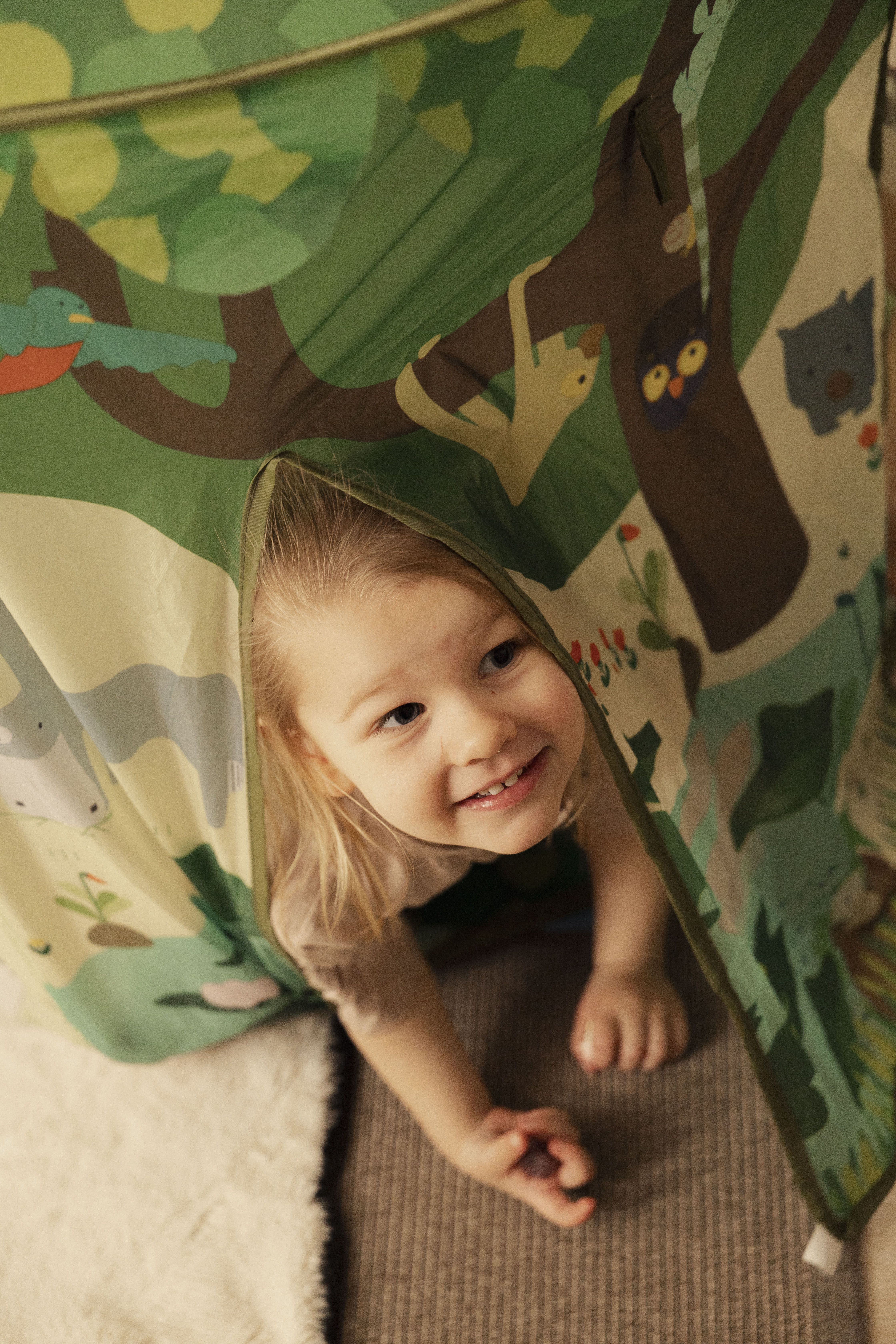 Smiling toddler peeks from colorful jungle play tent during playtime.