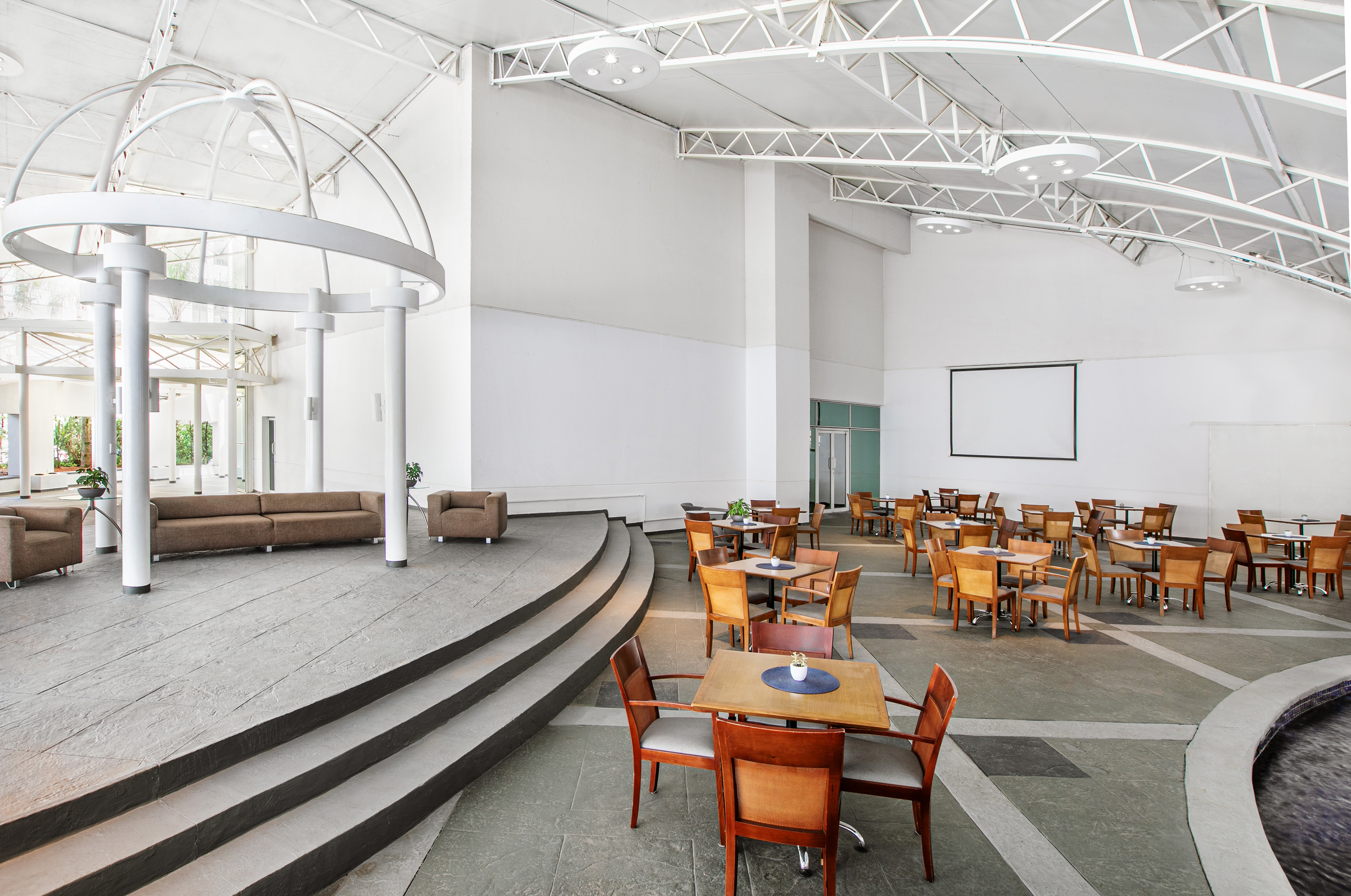 Modern event hall with lounge, dining tables, projector screen, and high ceiling.