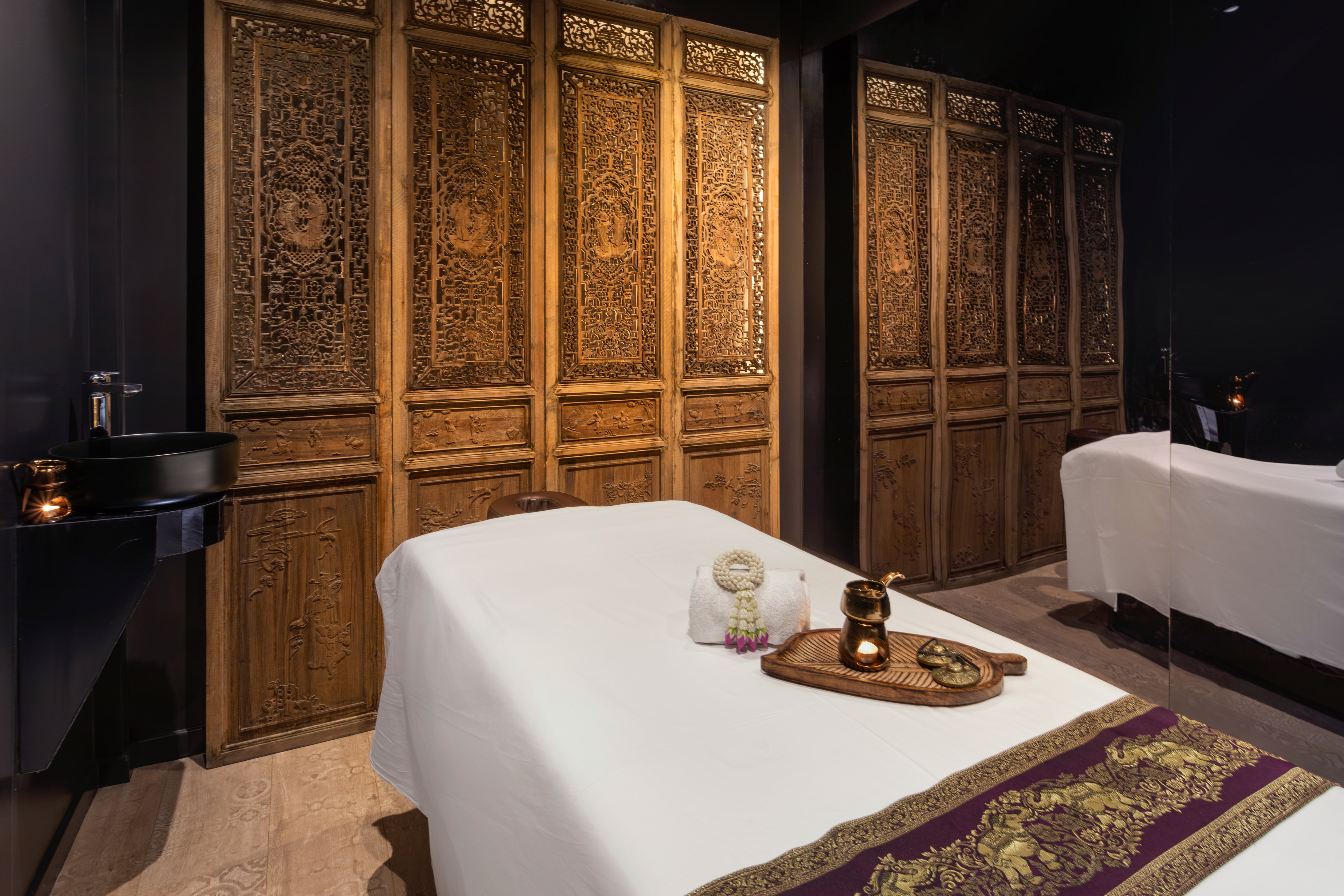 Thai spa room with ornate carved wood screens, massage bed, and relaxation decor.