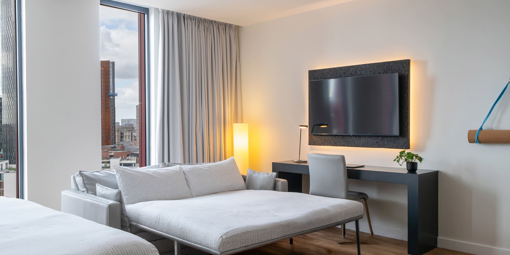 Modern hotel room with sofa bed, desk, wall-mounted TV, and city view.