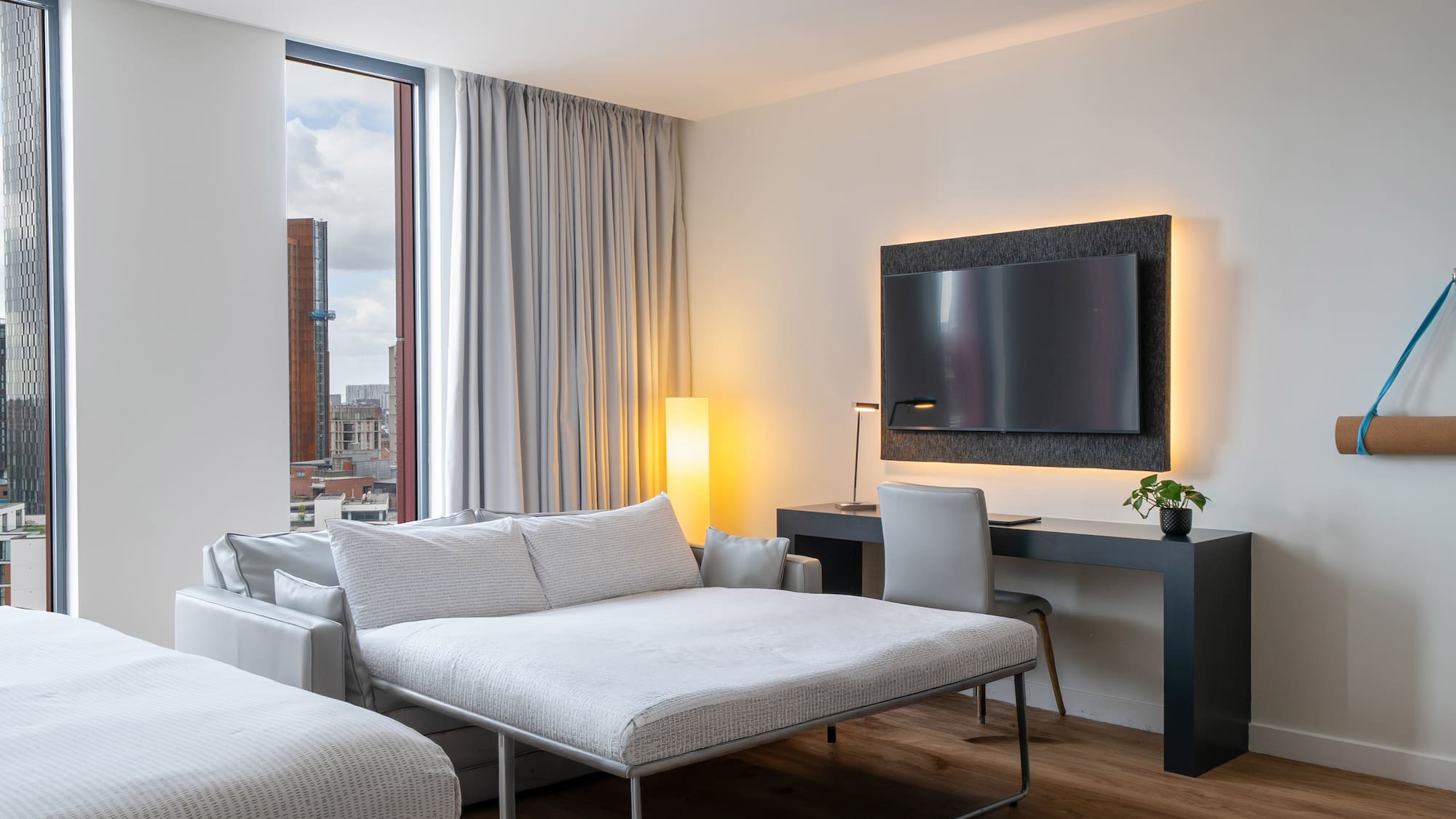 Modern hotel room with sofa bed, desk, wall-mounted TV, and city view.