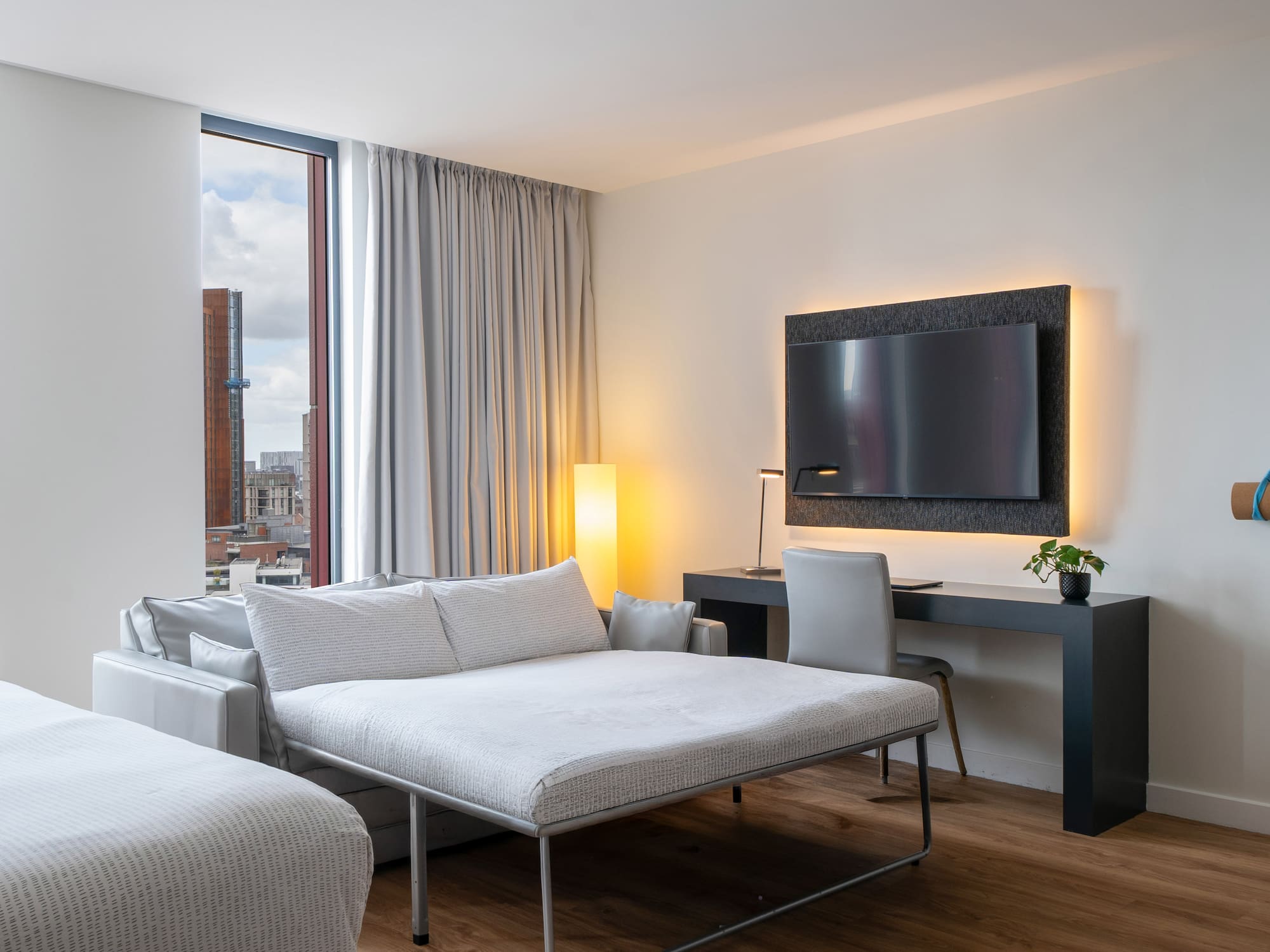Modern hotel suite with pull-out sofa bed, city view, desk, and wall-mounted TV.