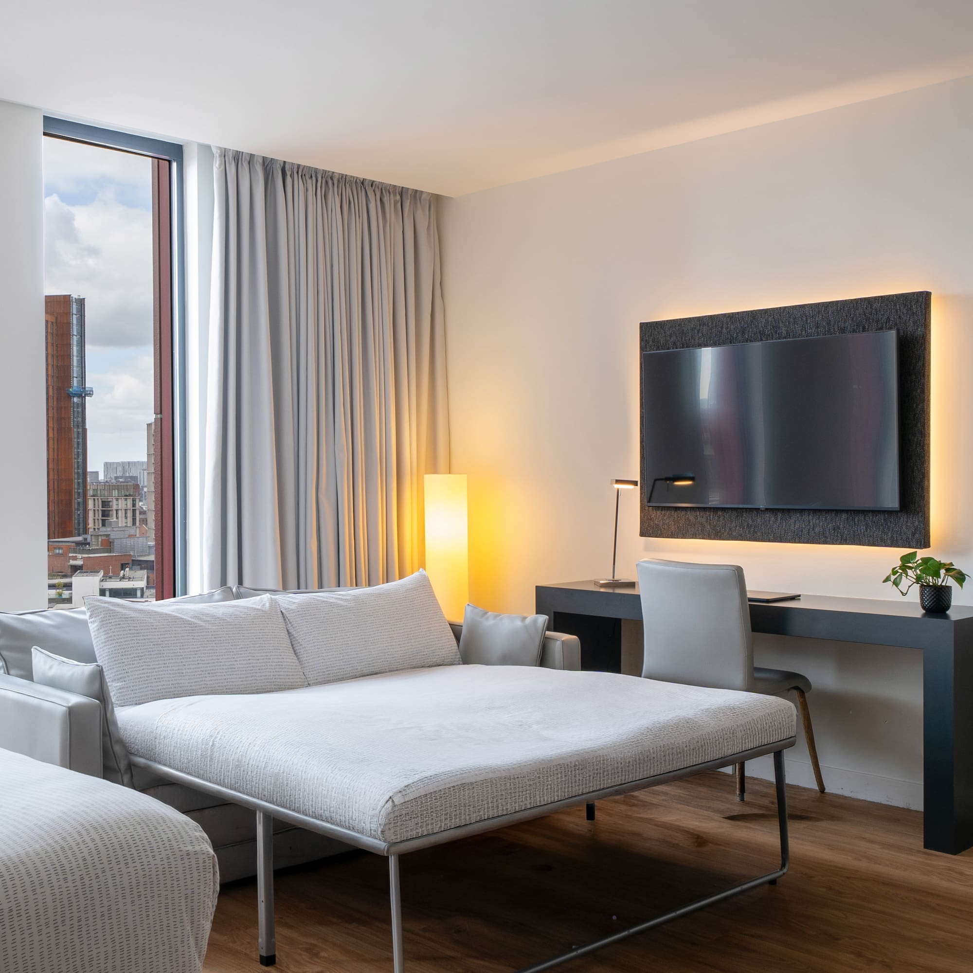 Modern hotel room with sofa bed, desk, wall-mounted TV, and city view.