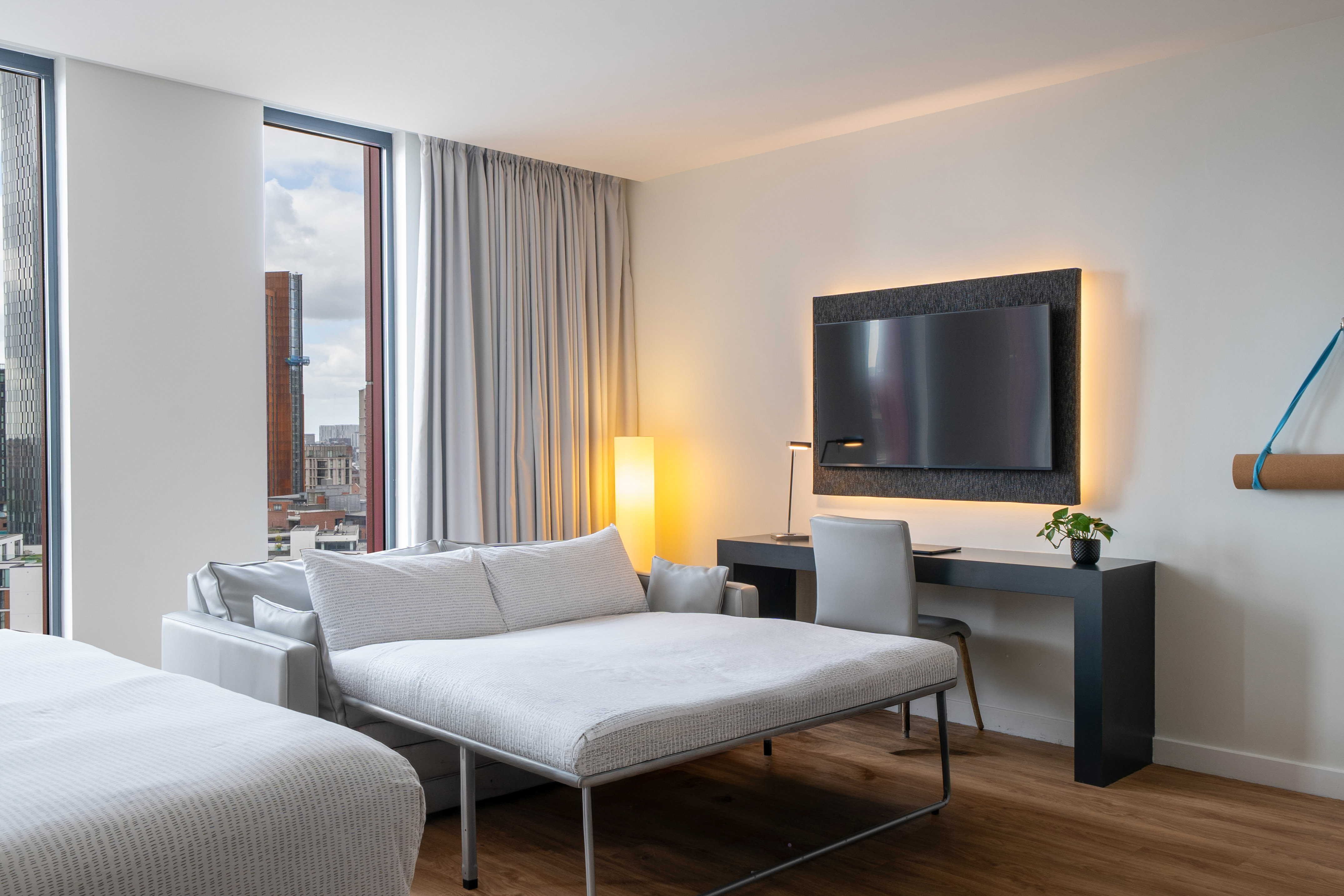 Modern hotel suite with pull-out sofa bed, city view, desk, and wall-mounted TV.