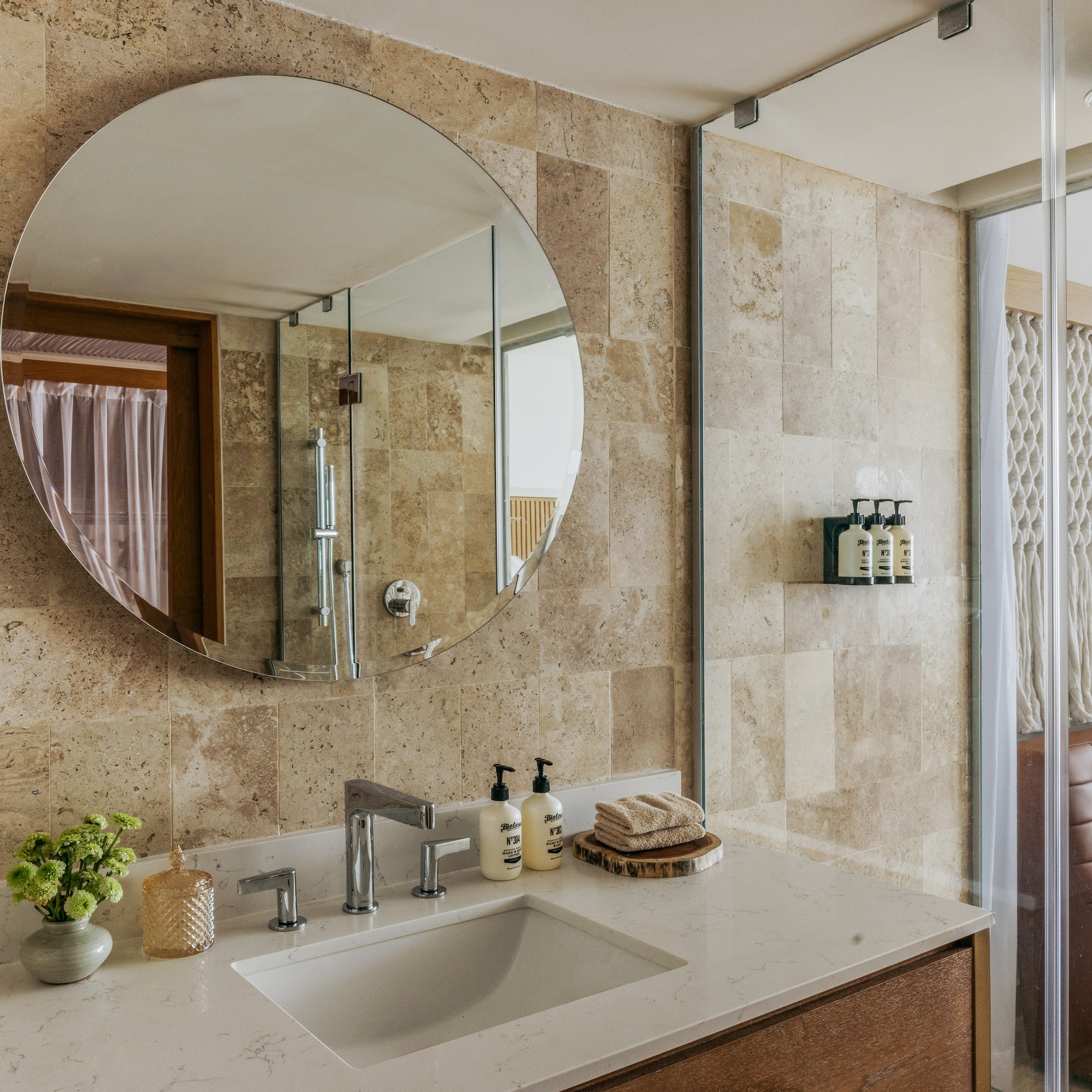Modern hotel bathroom with round mirror, travertine walls, and white vanity.
