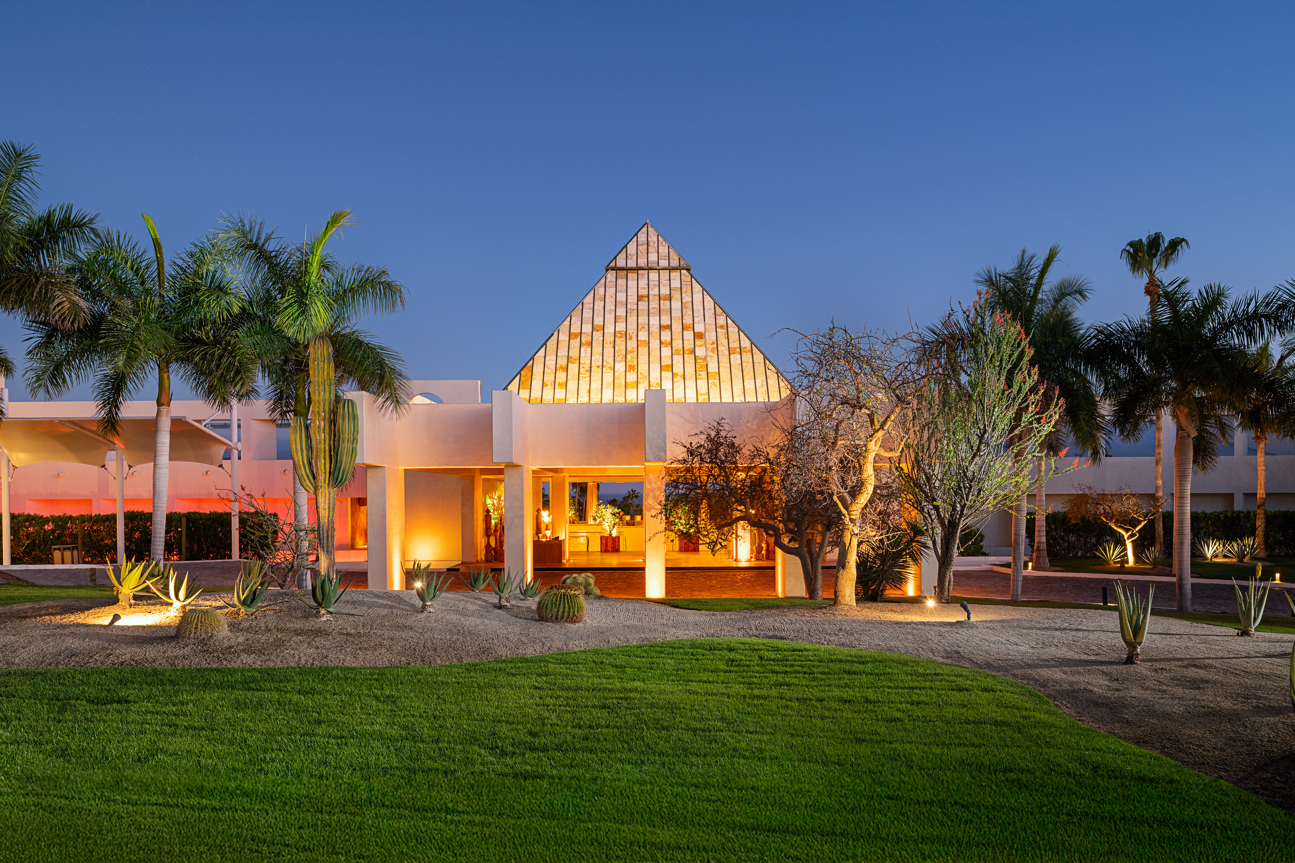 Pyramid-roofed resort facade at night with illuminated desert landscaping and palm trees.