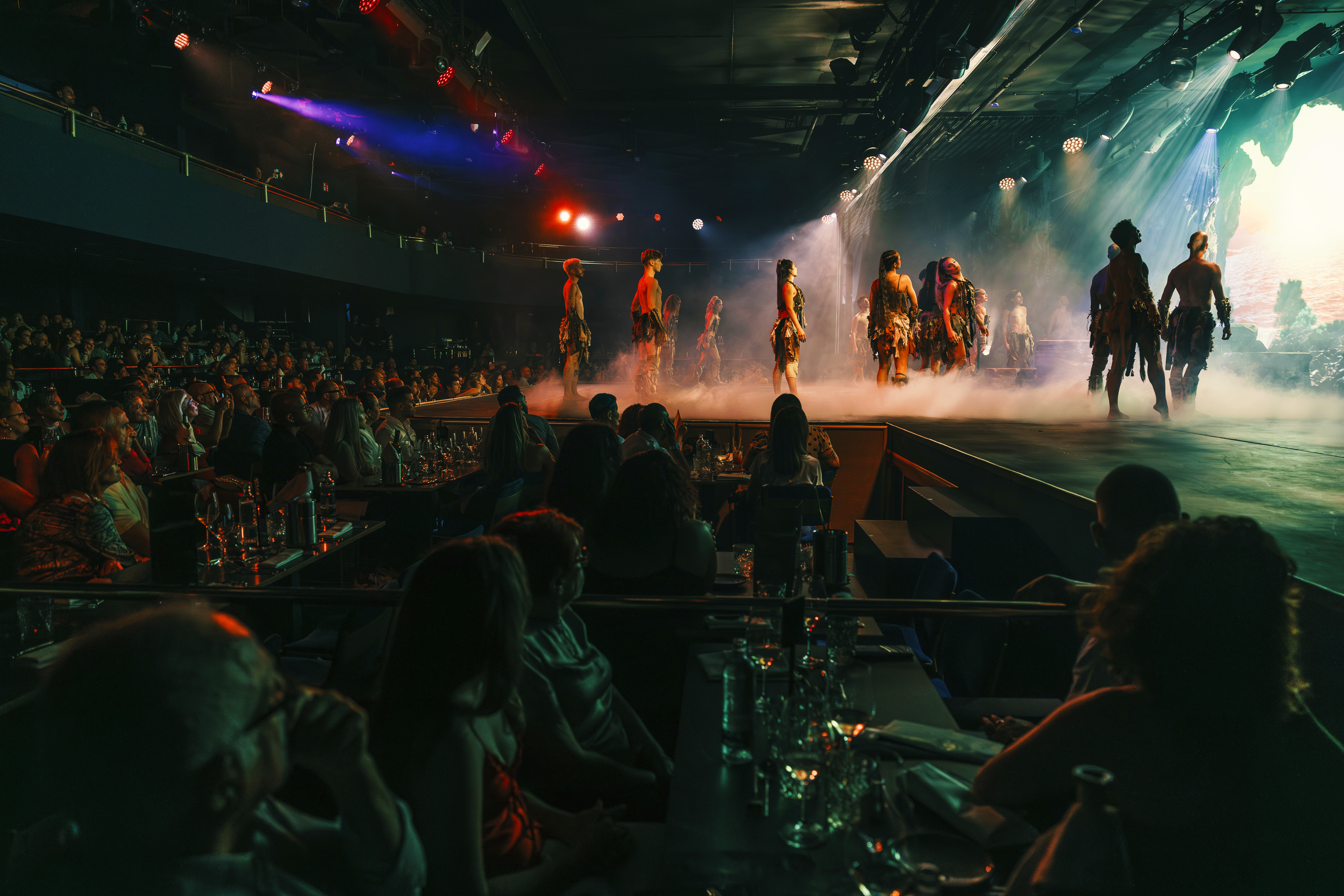 Audience views tribal dancers performing on a smoky stage with colorful lights.