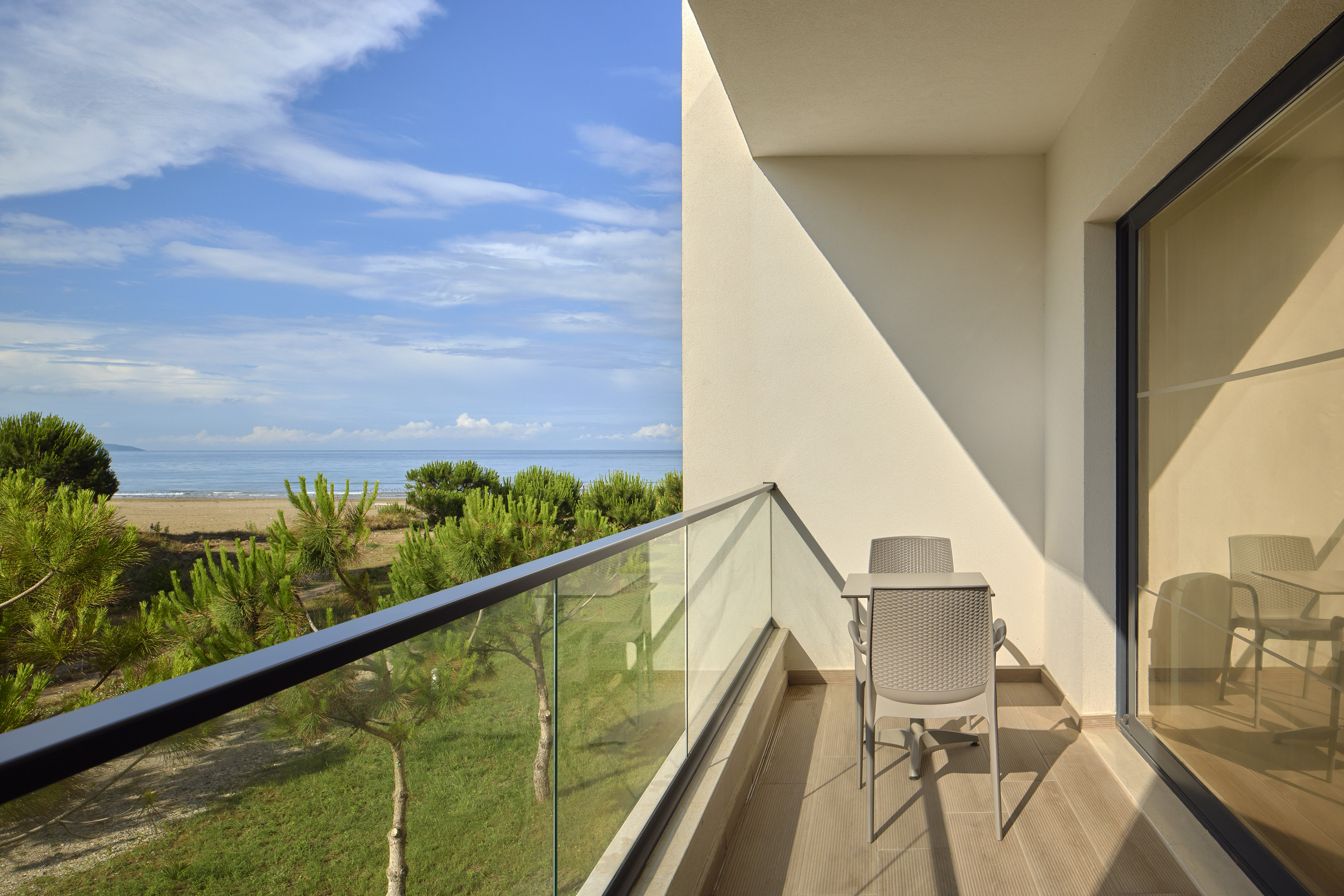 Coastal resort balcony with seating, beach, ocean, and lush trees under blue sky.