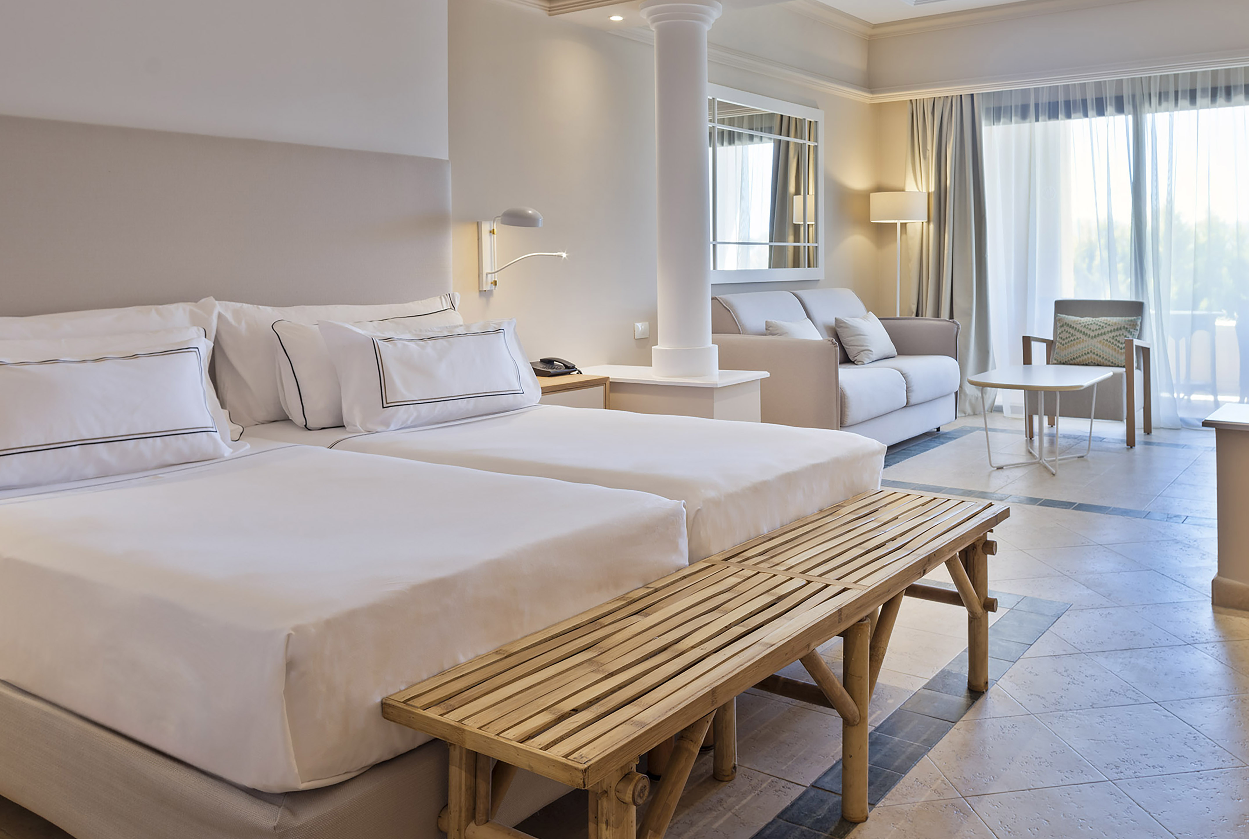 Modern hotel room with twin beds, sofa, bamboo bench, and bright windows.