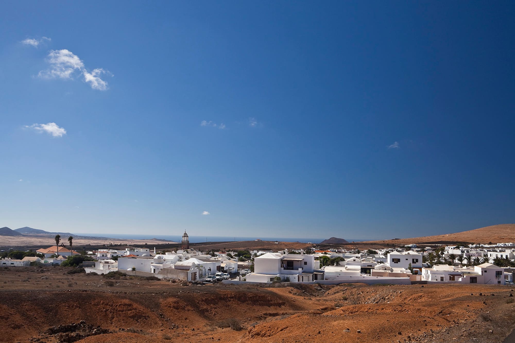 Pueblo blanco costero, paisaje árido rojizo, cielo azul y mar de fondo.
