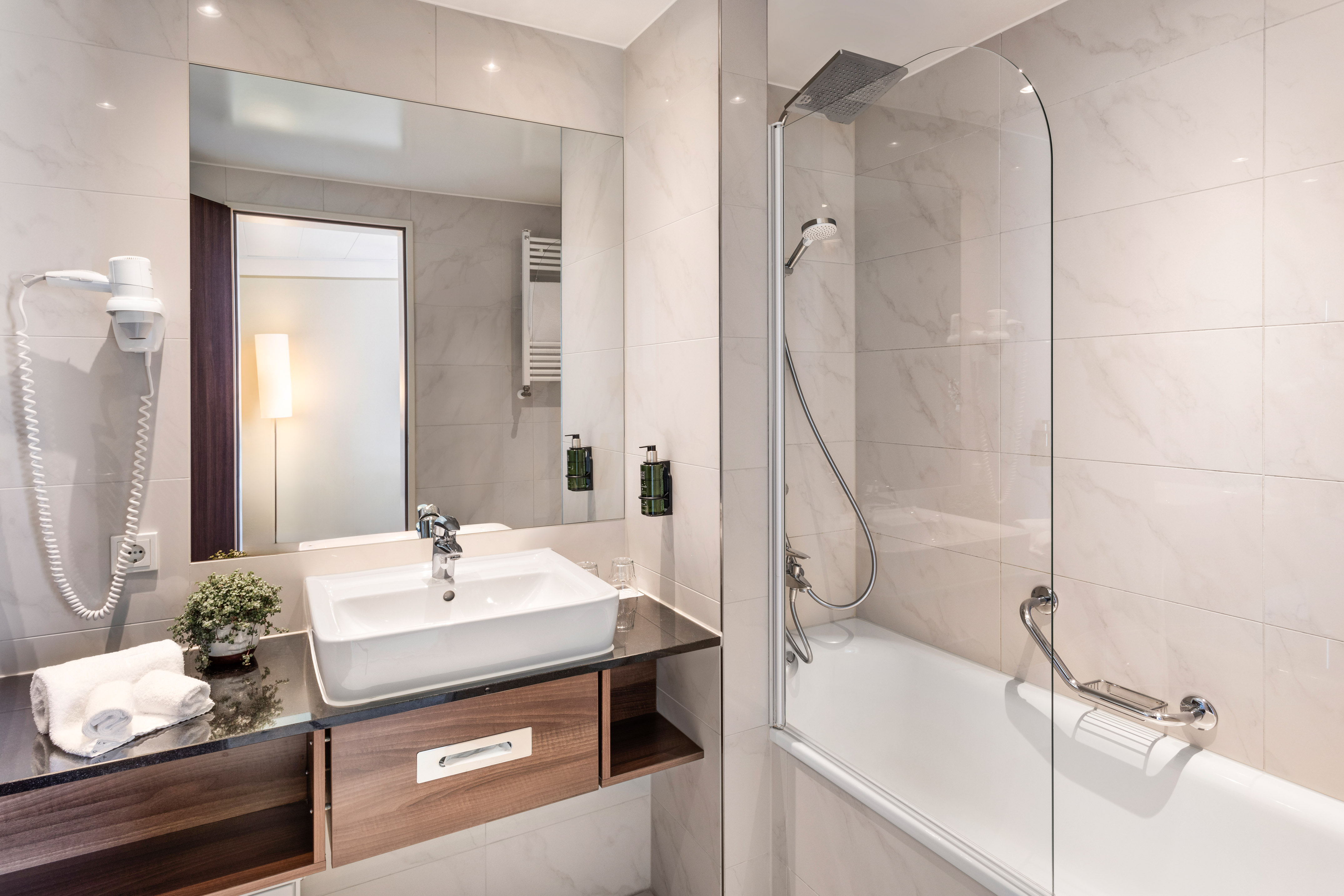 Modern hotel bathroom with white tiles, vanity sink, mirror, and shower-bathtub.