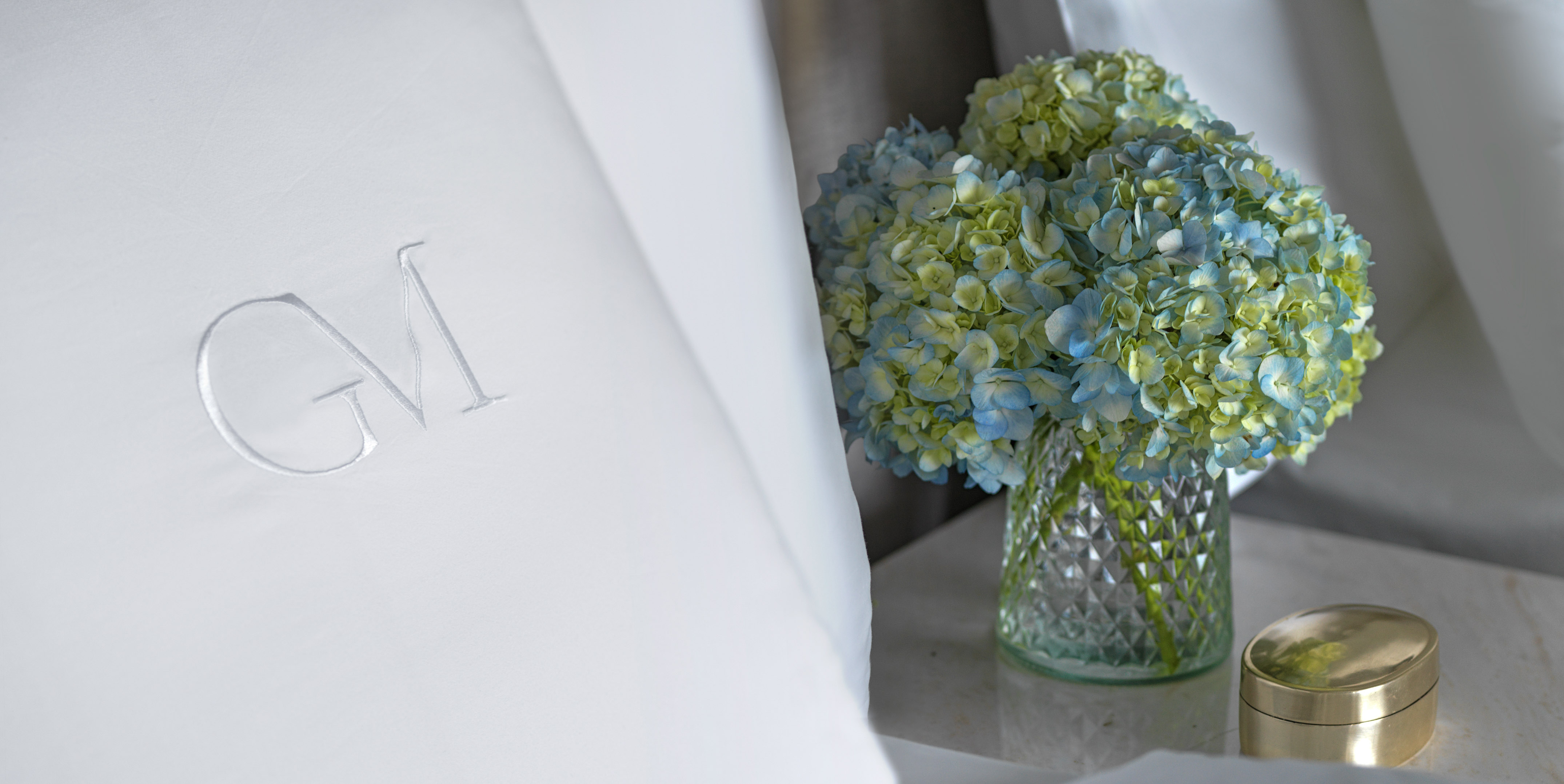 White monogrammed GM bed linen, blue hydrangeas in vase, and gold box.