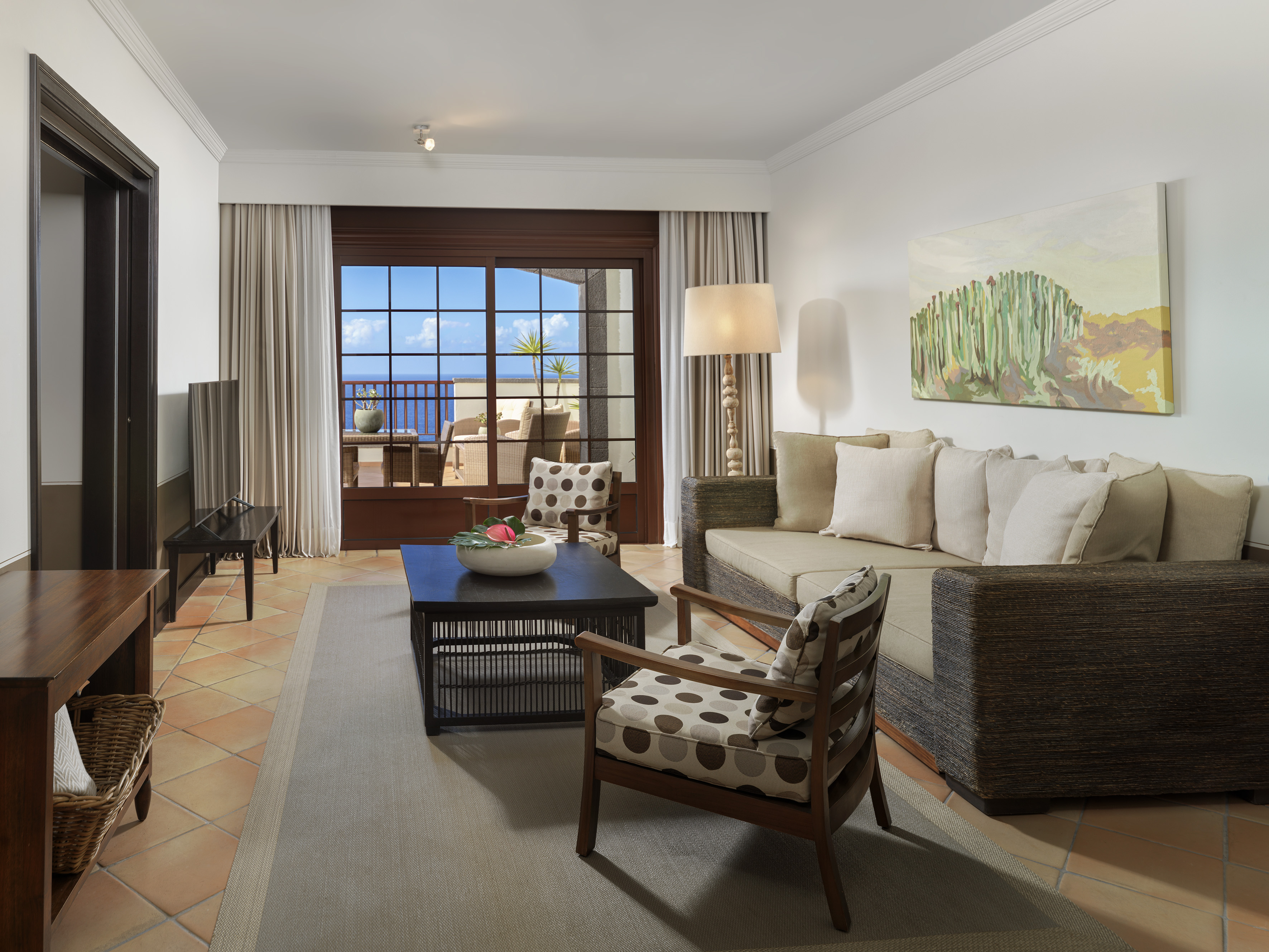 Resort living room with ocean view balcony, sofa, armchair, and cactus painting.