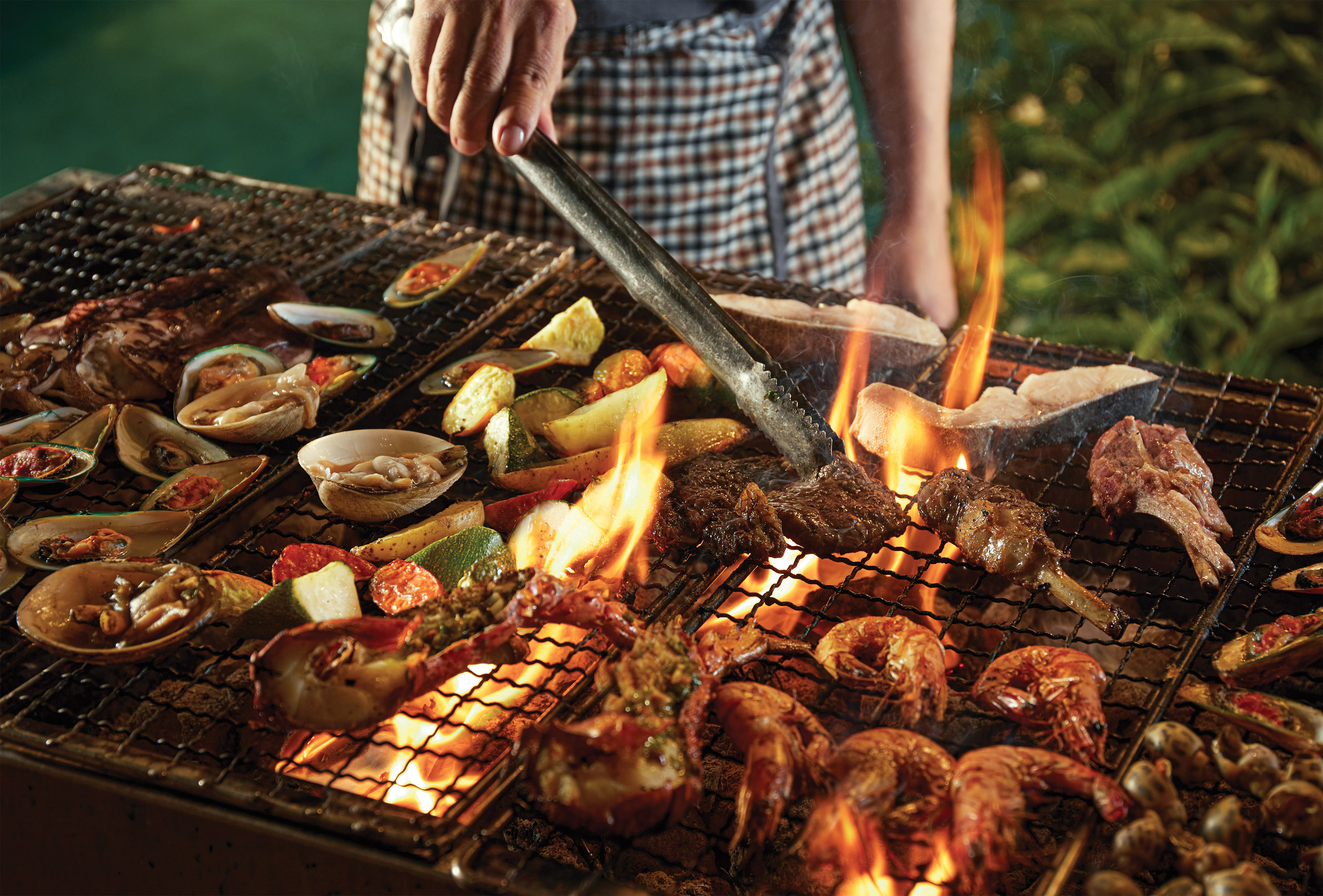 Outdoor BBQ with diverse seafood, meats, and vegetables grilling over flames.
